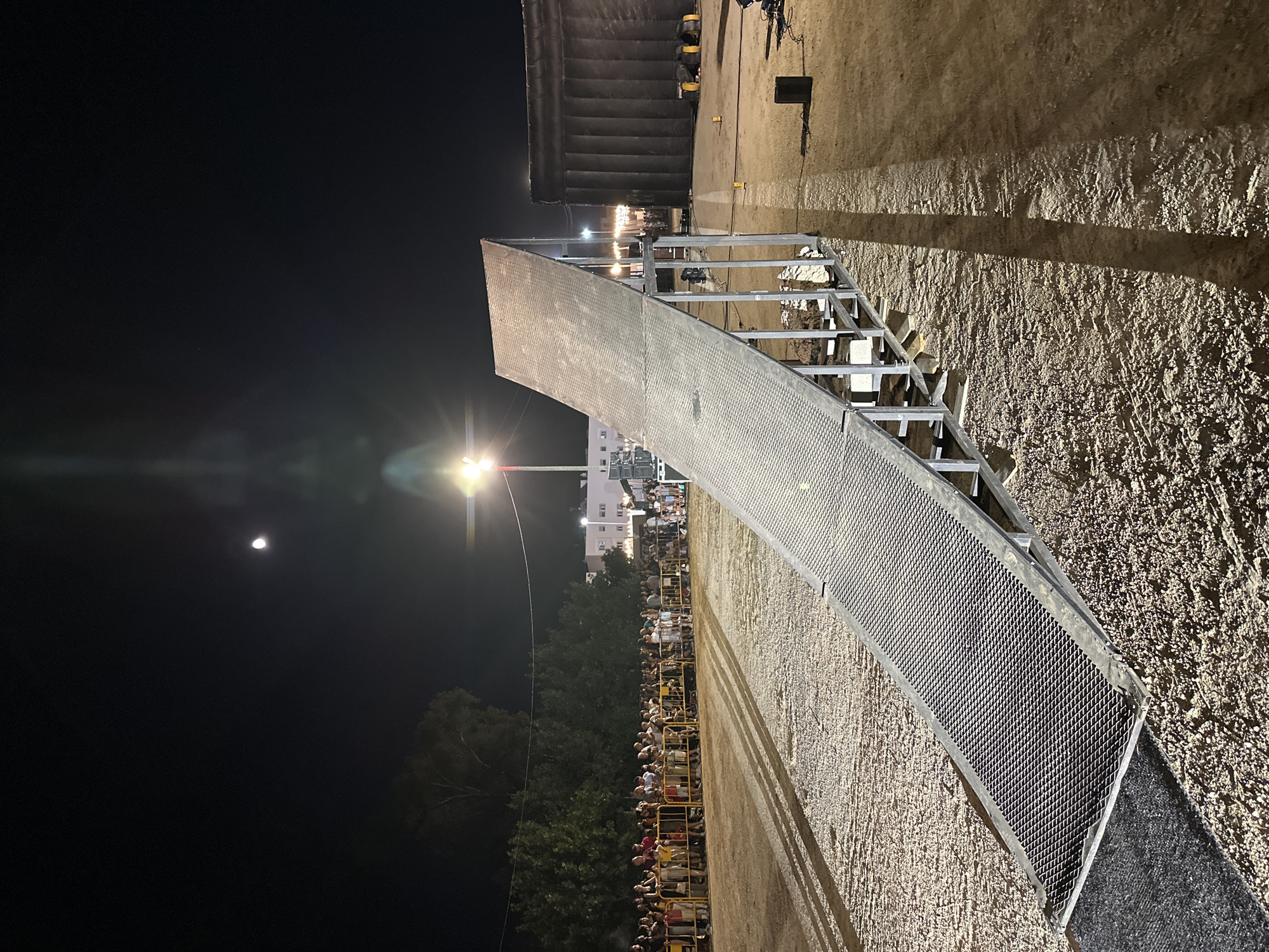 A long, metallic ramp stretches across a dirt-covered area at night, under a partially obscured moon and artificial lighting. The ramp, supported by a metal frame, has a textured surface and is angled upwards. A crowd of spectators, illuminated by lights, stands behind yellow barriers in the background, observing the scene. Adjacent to the ramp, a large, dark, inflatable structure is visible, as well as various pieces of equipment and lighting apparatus. The overall environment suggests an outdoor event or performance. The location is identified as San José del Valle, Spain.