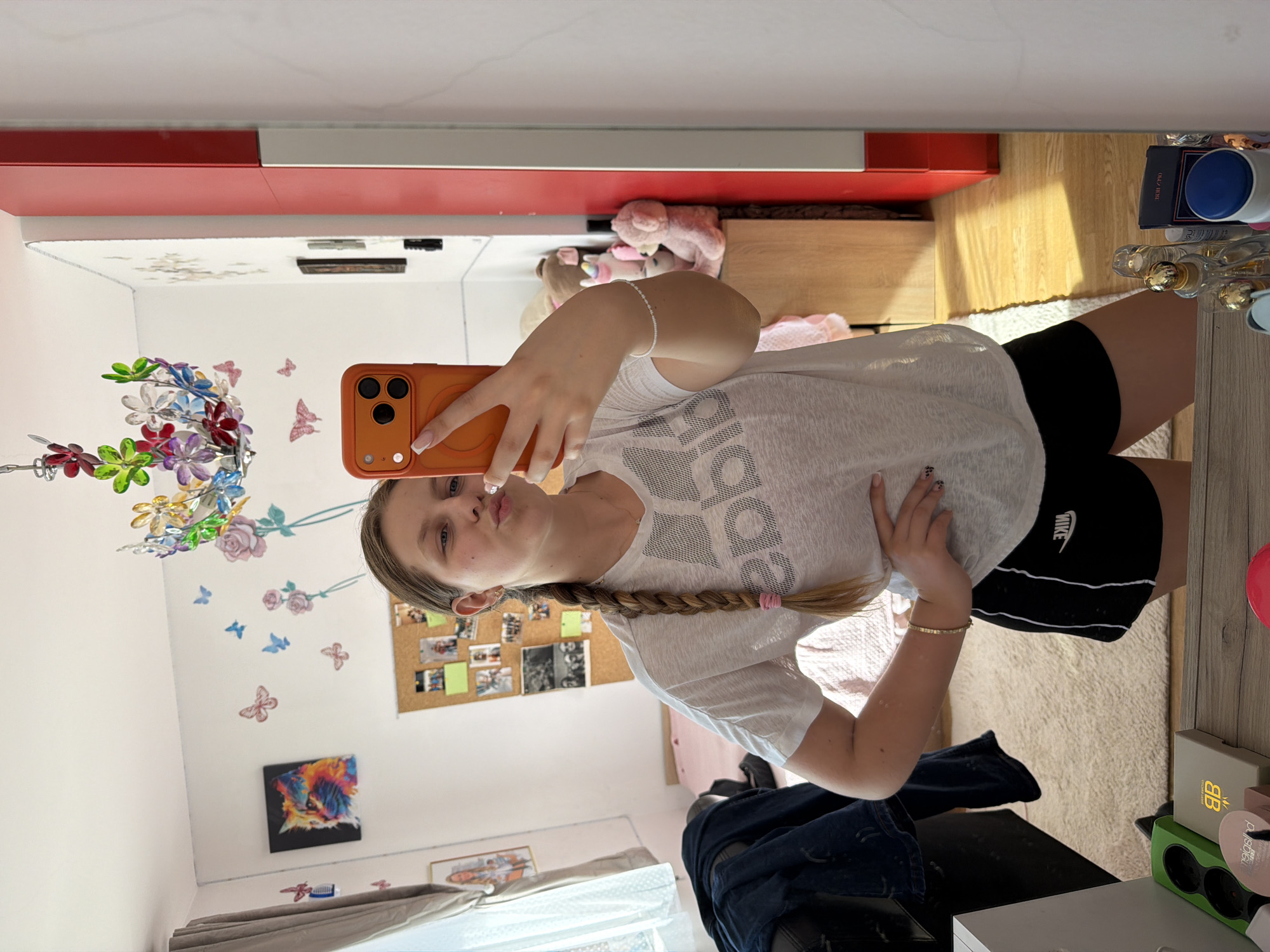 A young person is taking a mirror selfie in a bedroom. They are wearing a white t-shirt with an Adidas logo and black Nike shorts. They have a braid in their hair and are puckering their lips. The room is decorated with colorful butterfly decals and a painting of a cat. There is a corkboard on the wall with photos and a large mirror that reflects the room. In the reflection, a red cabinet and a wooden dresser with stuffed animals can be seen. The floor is carpeted and wooden. The person is holding an orange smartphone.