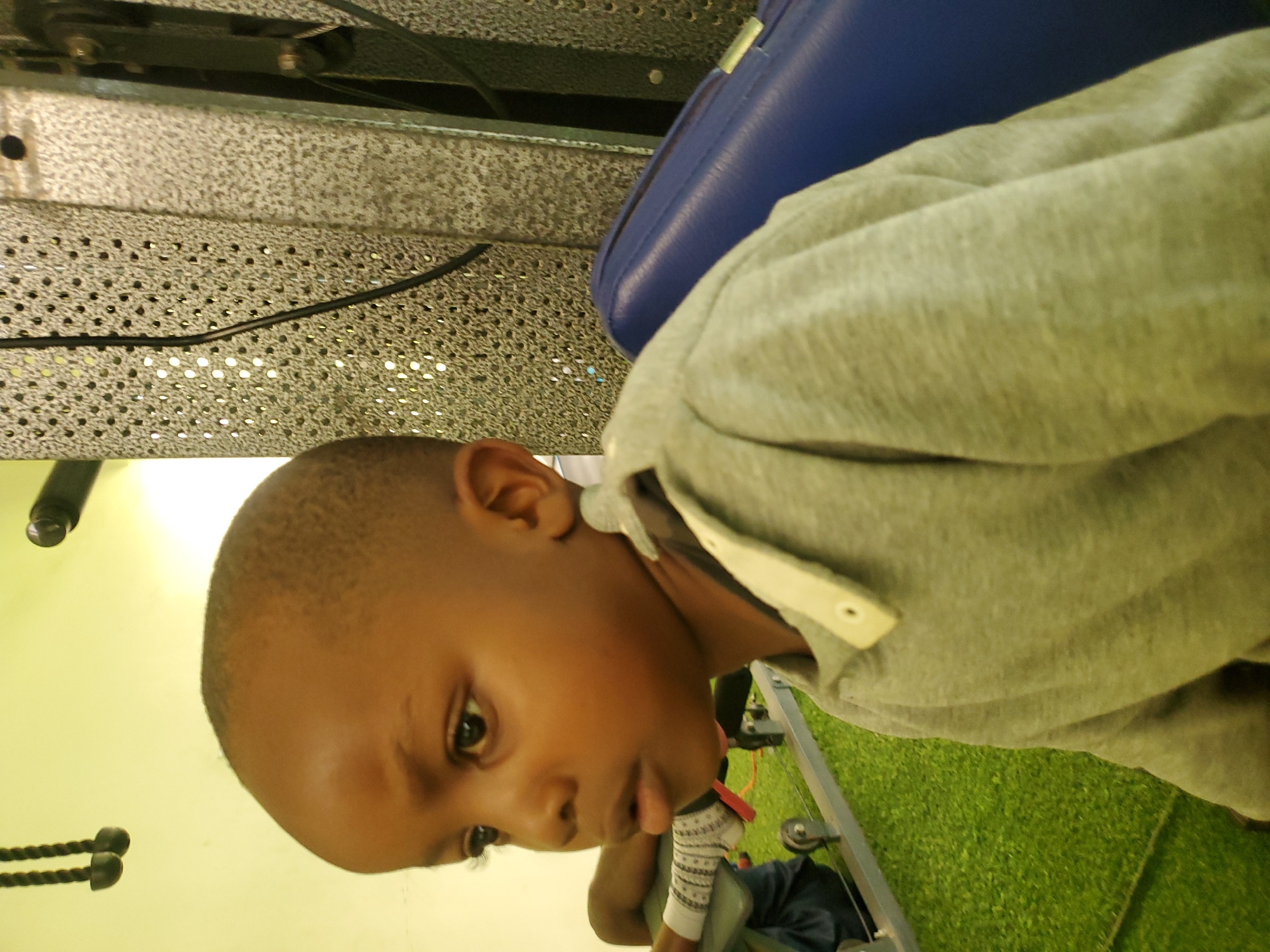 A medium shot captures a young boy with dark skin and a closely cropped haircut, positioned in the foreground to the left. He is looking to his left, with his mouth slightly open and his gaze directed downwards. He is wearing a gray long-sleeved hooded sweatshirt with a light-colored collar and buttons. Behind him, to the left, a person's legs are visible, wearing white socks with a pattern of gray and white stripes. The background features a green, artificial turf surface. To the right of the boy, a portion of a blue padded object is visible, possibly a seat or cushion. Further in the background, a metallic, perforated wall is observed, characteristic of gym equipment. A black cable hangs vertically down the perforated wall. The overall setting appears to be an indoor recreational facility, possibly a gym. The lighting is bright, with a slight yellow tint. The image was taken in Jos, Nigeria.