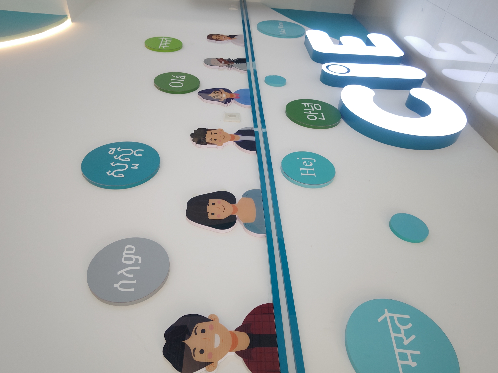 The image depicts a wall decoration in a building in the Chongqing High-tech Zone, China. The decoration features the word "LIFE" in large, illuminated white letters against a blue background. Along the wall are various cartoon images of people, with corresponding circular plaques displaying the word "hello" in different languages. The plaques are arranged along a blue line that runs parallel to the "LIFE" letters. The scene is well-lit.