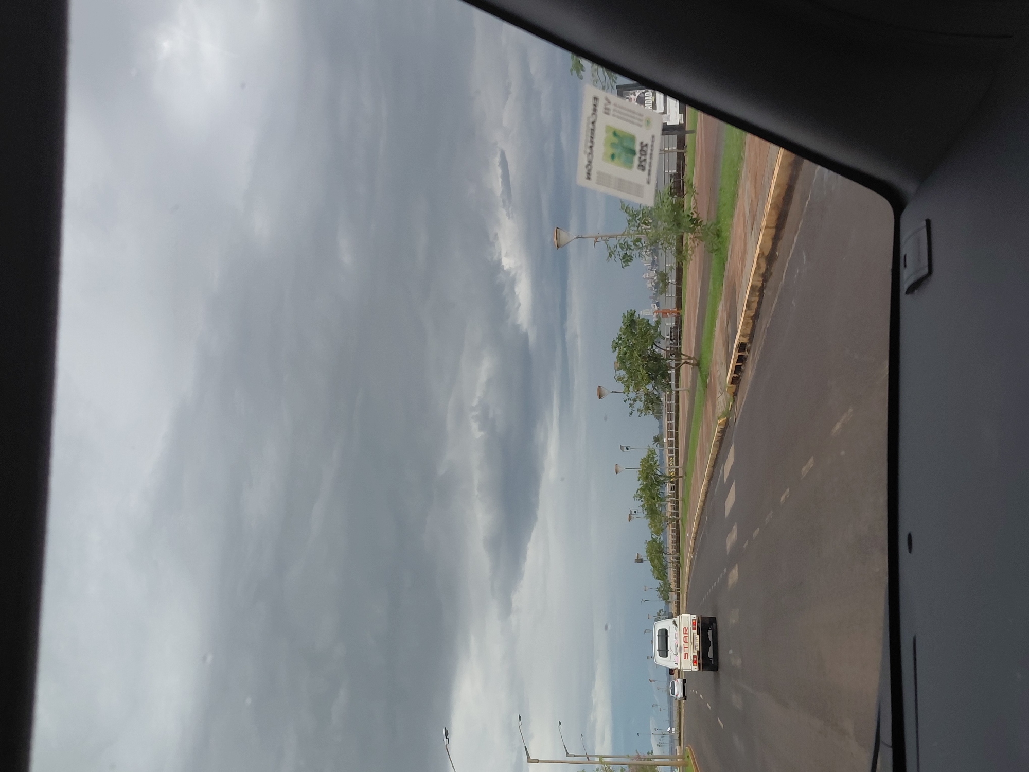 The image shows a view from inside a vehicle looking out at a cloudy sky and a road with a waterfront promenade. A white pickup truck with "STAR" branding on the back is driving on the road. A white car is visible further ahead. The promenade has streetlights and trees. A sticker with green graphics and text, appearing to be an inspection sticker, is affixed to the vehicle's windshield on the passenger side. In the background, a city skyline is visible across the water. The location is the city of Encarnación, Paraguay, specifically along the Costanera, the city's waterfront avenue.