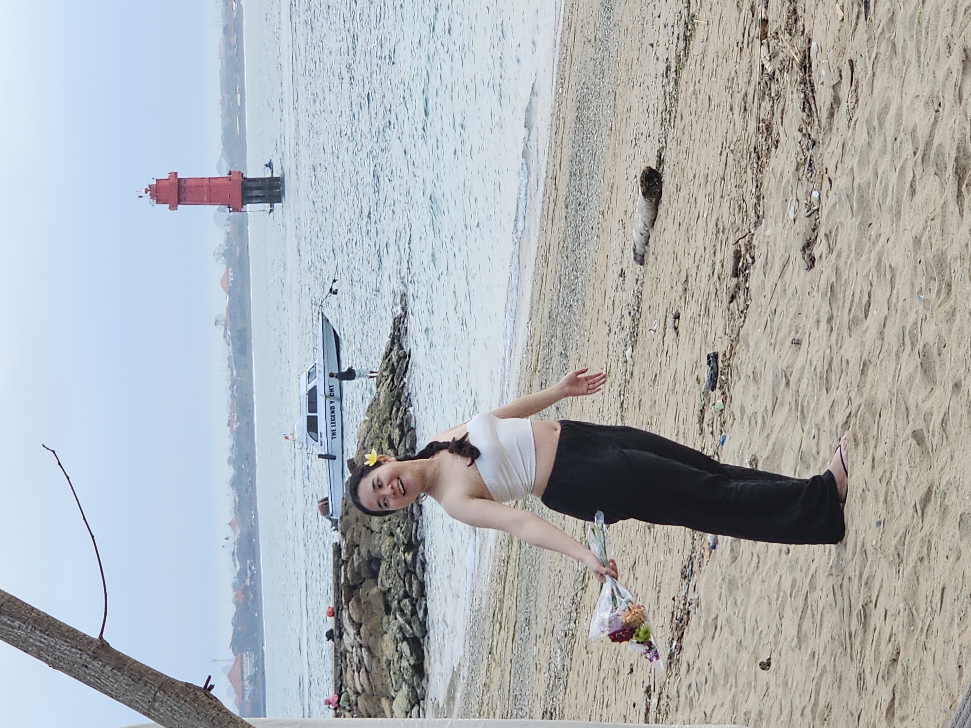A young woman with a yellow flower in her hair stands on a sandy beach, holding a bouquet of flowers. She is wearing a white tube top and black pants. Behind her, the ocean stretches out, with a red lighthouse visible in the distance. A boat with "THE LEGEND YACHT" written on its side is also present in the water. The beach is lined with rocks and debris. The sky is overcast. This scene appears to be at Sanur Beach in Denpasar, Indonesia, given the prominent red lighthouse, which is a well-known landmark there.
