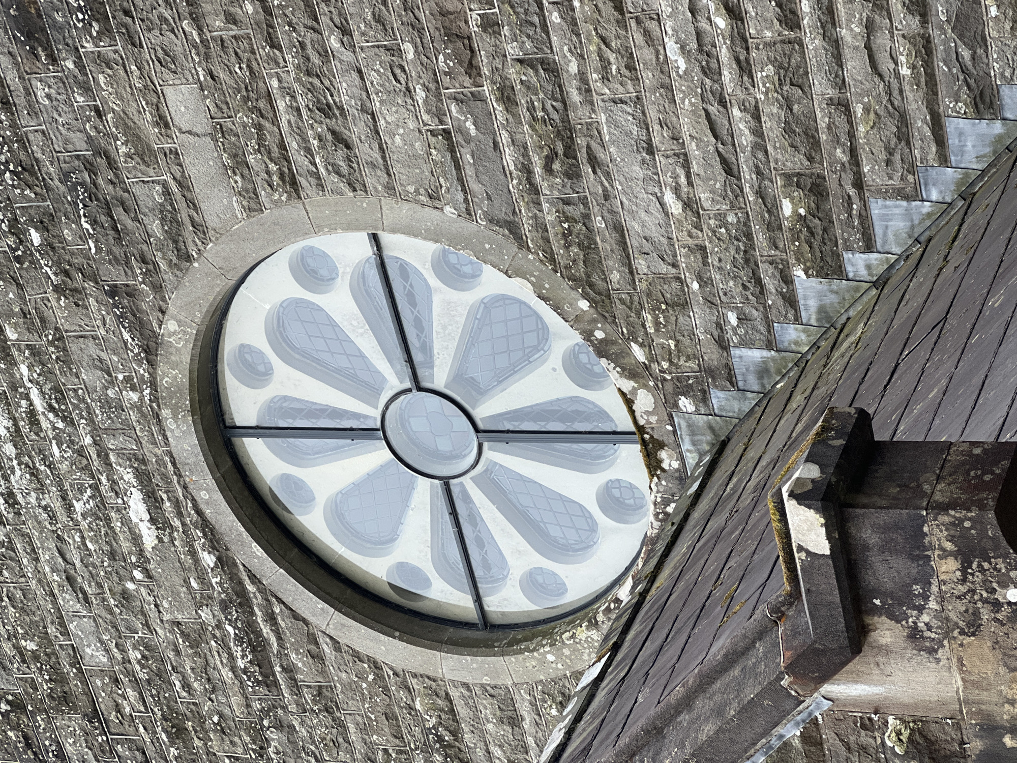 This is a top-down, close-up shot of the exterior of a stone building, likely a church or historical structure, given the architectural details. The primary subject is a large, circular stained-glass window, often referred to as a rose window, which is stylized with a floral or petal-like design. The window is framed by rough-hewn grey stone blocks that form the wall.
Adjacent to the window, and dominating the right side of the frame, is a dark-tiled roof, sloping downwards and away from the viewer. The roof tiles appear to be slate and are weathered, with patches of moss and lichen visible, suggesting an older structure. The stone work of the wall is also weathered, with white and green patches of lichen scattered across its surface, indicating exposure to the elements over time.
The stonework is laid in horizontal courses, with individual blocks having a rough texture and visible mortar lines. The window itself is a prominent feature, with a central circular pane surrounded by radiating, petal-shaped panes of glass. These panes are a light, translucent grey with a subtle pattern etched into them, possibly representing leaded glasswork or a frosted effect. The glass has a reflective quality, showing some of the surrounding architectural elements.
The scene is lit by natural light, casting subtle shadows that highlight the textures of the stone and the roof tiles. The time of day appears to be daytime, likely morning or afternoon, given the brightness and the angle of the light. There are no people visible in the frame, and the focus is entirely on the architectural elements. The overall impression is one of age, solidity, and a sense of place, consistent with historical buildings found in locations like Mountcharles, Ireland. No visible text is present in the image.