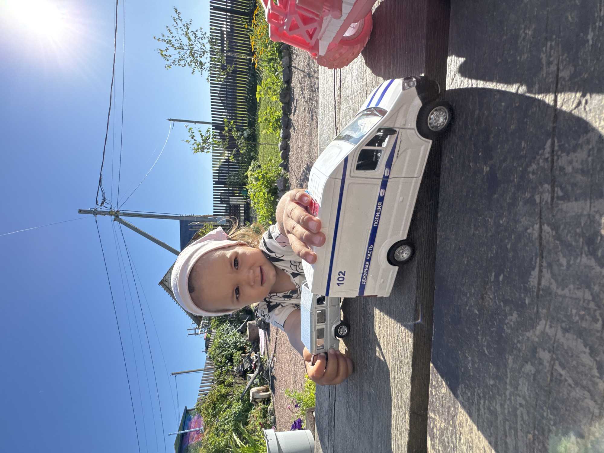 A young girl is playing with toy vehicles outdoors on a sunny day. The girl, wearing a light-colored headscarf and a patterned t-shirt, is seated at a wooden table. She is holding a silver toy car in her left hand and reaching towards a white toy police van with blue stripes. The police van has "102" and "ДЕЖУРНАЯ ЧАСТЬ ПОЛИЦИЯ" written on its side. A pink toy vehicle is partially visible to the right of the police van. The background shows a wooden fence, green foliage, and a clear blue sky with the sun shining brightly. The location is identified as Kempelevo, Russia.