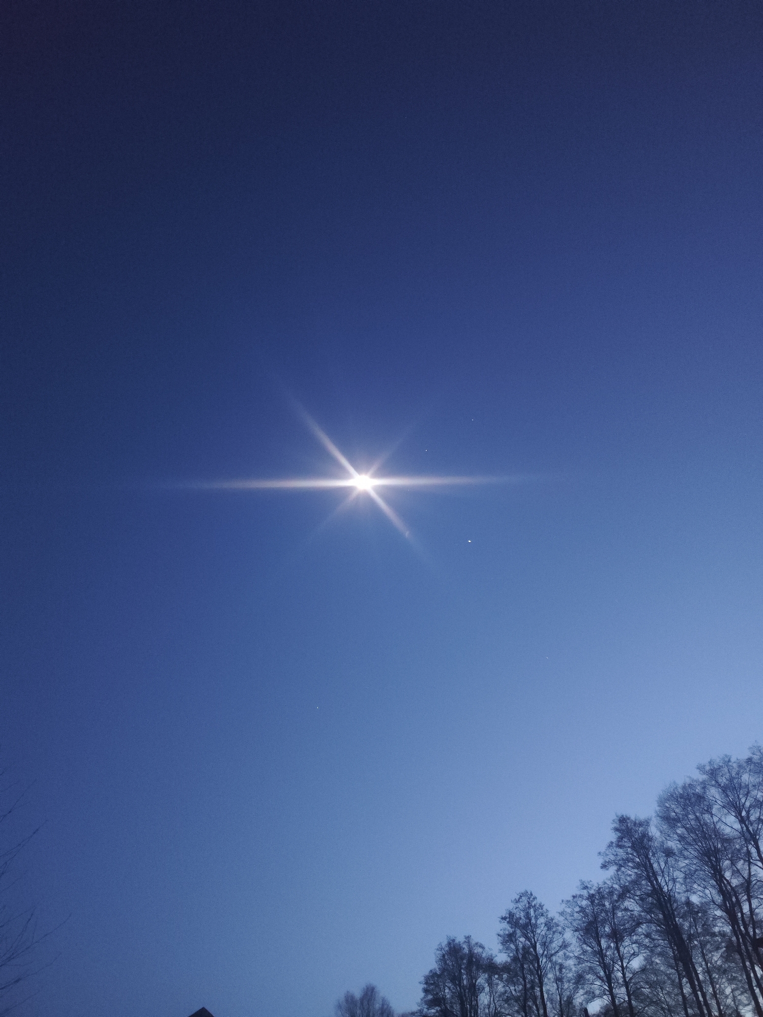 The image displays a deep blue sky dominated by a remarkably bright, star-like celestial object positioned in the upper center. This object exhibits a prominent lens flare, casting radiant light horizontally and vertically across the sky. To the lower right of this intense light source, a smaller, dimmer celestial body is visible, and several tiny specks, possibly distant stars, are scattered across the dark blue expanse.
The sky shows a gradient, transitioning from a darker, richer blue at the top to a slightly lighter shade towards the lower right horizon. This coloration, combined with the visible celestial bodies, suggests either a clear twilight period (dawn or dusk) or a very clear night with a prominent planetary or stellar object. The weather appears clear, with no clouds.
In the lower right corner, the intricate, leafless branches of bare deciduous trees are silhouetted against the lighter sky, indicating late autumn, winter, or early spring. A faint, dark silhouette of what appears to be a building's roofline is barely visible at the very bottom left edge of the frame. The scene evokes a sense of calm and vastness. No text is visible in the image.
