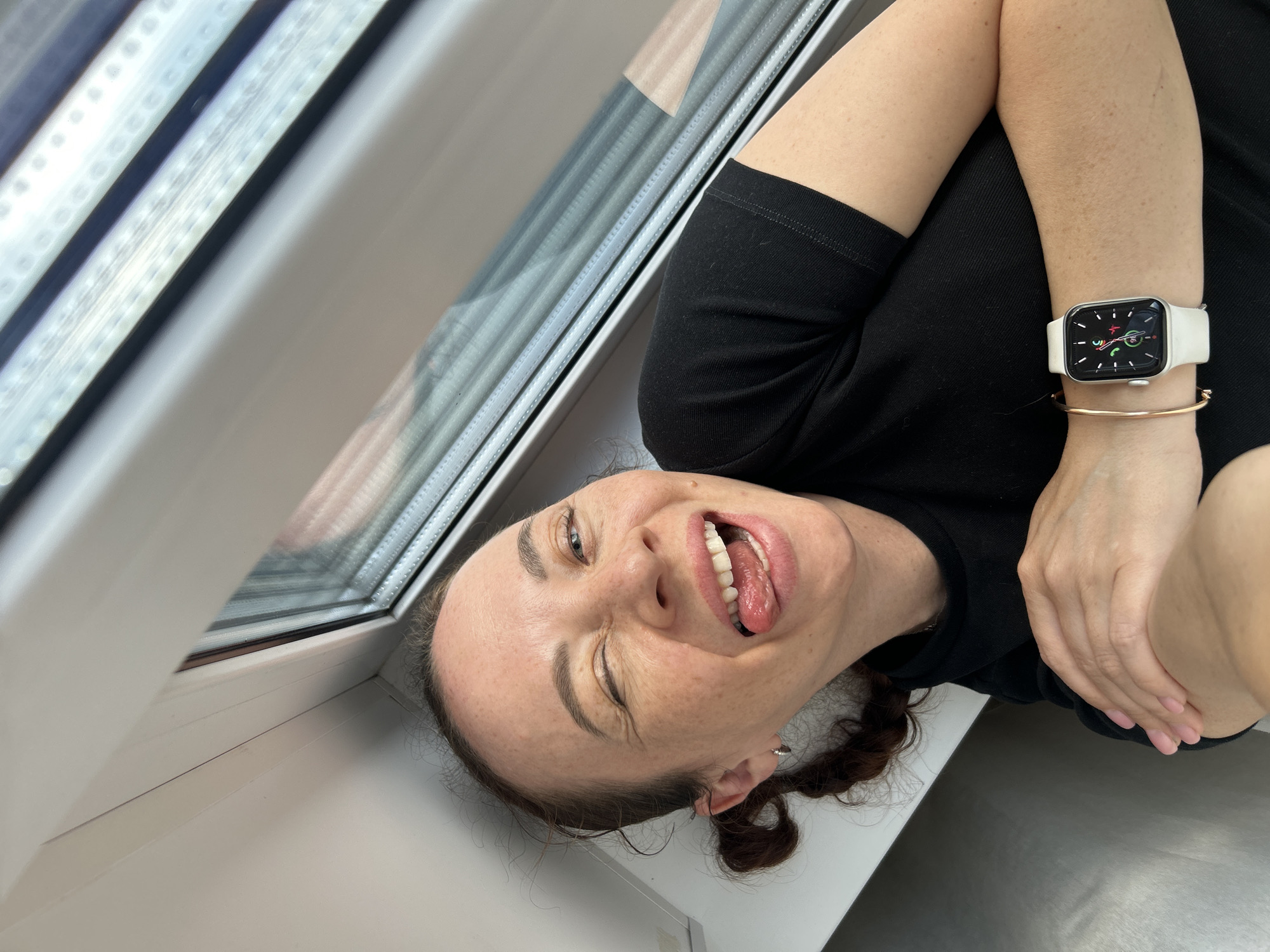 A woman with light brown hair pulled into two low pigtails is captured in a close-up, high-angle selfie. She is winking with her left eye and sticking out her tongue, with her mouth open. She is wearing a black t-shirt and a white Apple Watch with a rose gold bracelet. Her hands are crossed over her chest. The background consists of a white windowsill and a window with horizontal blinds, suggesting an indoor setting, possibly on a train or a room with a large window. No elements in the image suggest a specific location in Yekaterinburg, Russia.