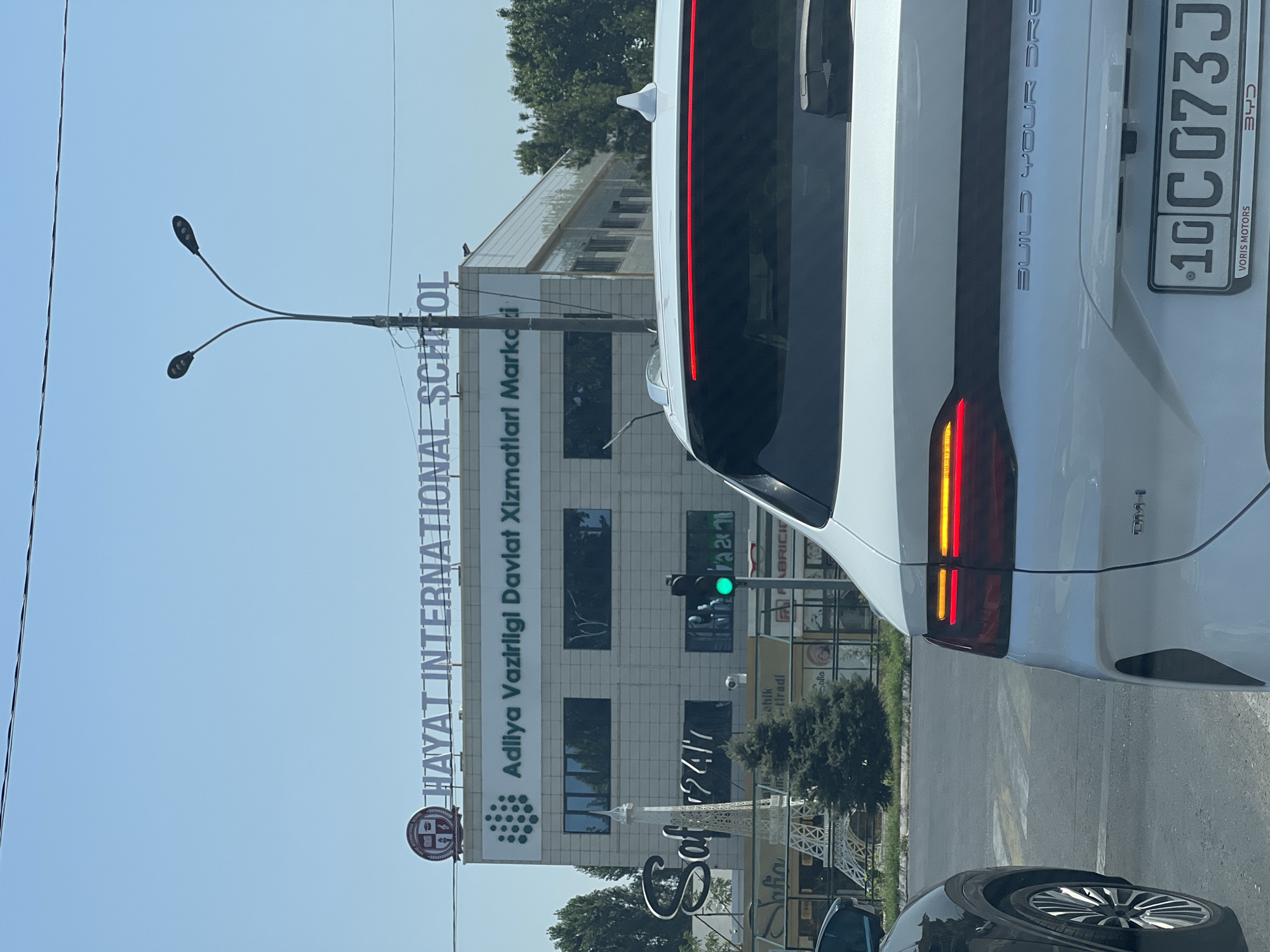 This is a photograph taken from inside a vehicle in Kibray, Uzbekistan. The image captures the rear of a white electric SUV, a partial view of a black car to its left, and the facade of a multi-story building in the background. The building is identified by prominent signage as "HAYAT INTERNATIONAL SCHOOL" and "Adliya Vazirligi Davlat Xizmatlari Markazi". A traffic light is visible, currently displaying a green signal. In front of the building, a decorative Eiffel Tower replica and several trees are present. The sky is clear and blue, indicating a sunny day. The license plate of the white SUV reads "10 C 073 J".