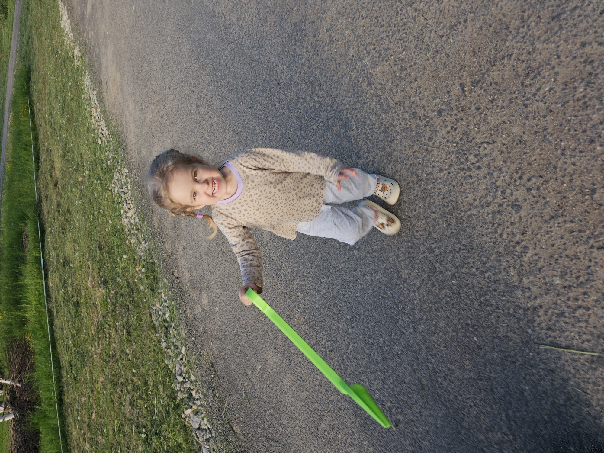 A girl lies on a paved road near a grassy area with trees. She is fair-skinned with blonde hair and is wearing a light-colored, patterned shirt and pants, along with printed shoes. She is smiling and holding a green plastic scoop-like toy. The road and grassy area suggest an outdoor, possibly park-like setting. The location of the photograph is Мышкино, Russia.