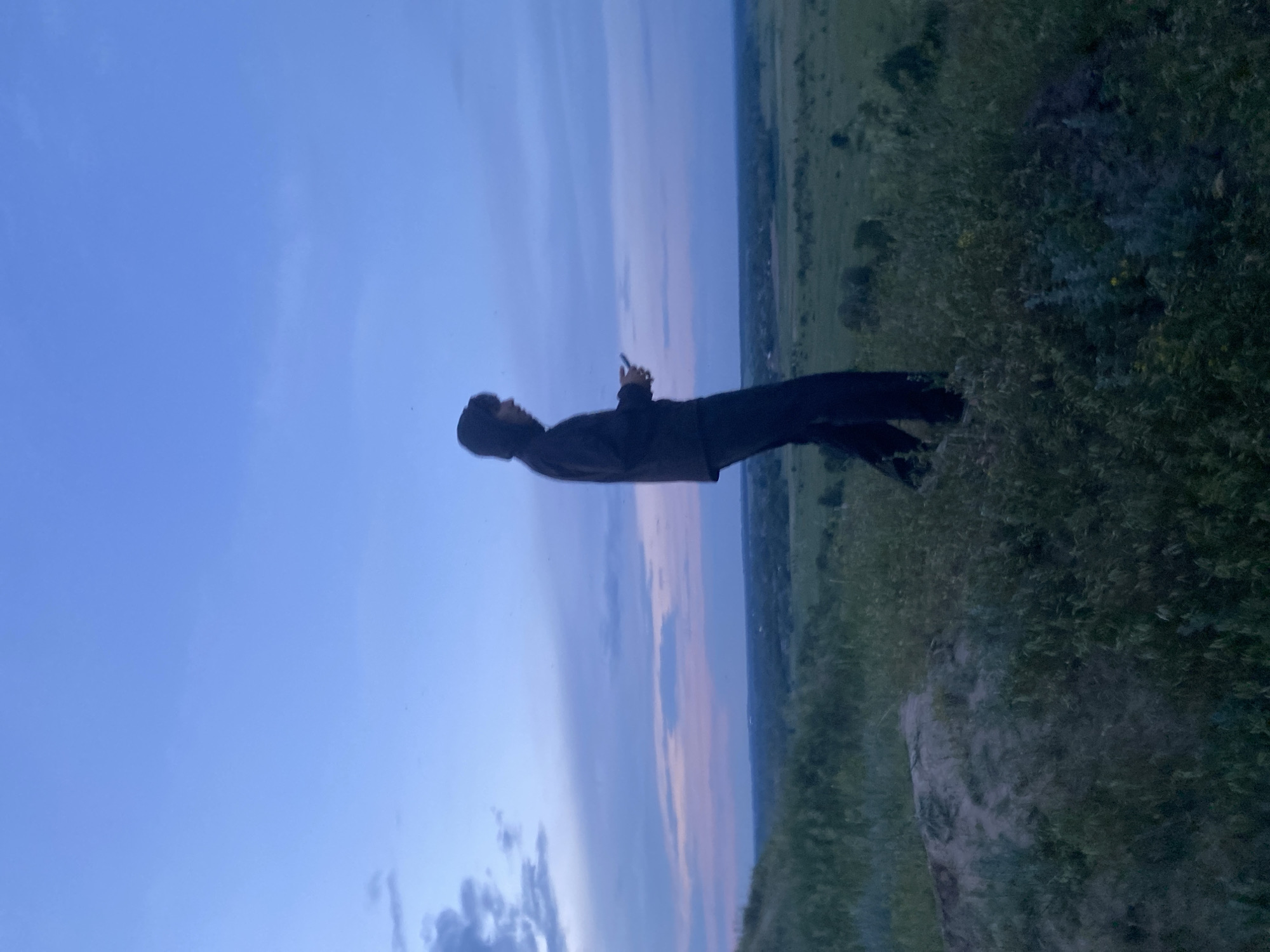 The image captures an outdoor scene at twilight near Ustyvytsia, Ukraine. A lone individual, depicted as a dark silhouette, stands on a grassy hill overlooking an expansive, flat rural landscape. The person is dressed in dark attire, including a hooded jacket and pants, and is positioned in profile, facing towards the right side of the frame. Both hands are engaged with a small, rectangular object, likely a smartphone, held at chest level. The immediate foreground features a hill with varying textures of green vegetation and patches of exposed earth. Beyond the hill, the midground and background reveal a broad, flat plain characterized by green fields, scattered trees, and distant, indistinct structures suggesting a settlement. The sky displays a gradient of colors, transitioning from deep blue at the top to lighter blues, purples, and faint pinks closer to the horizon, with several dark clouds present, primarily on the left side.