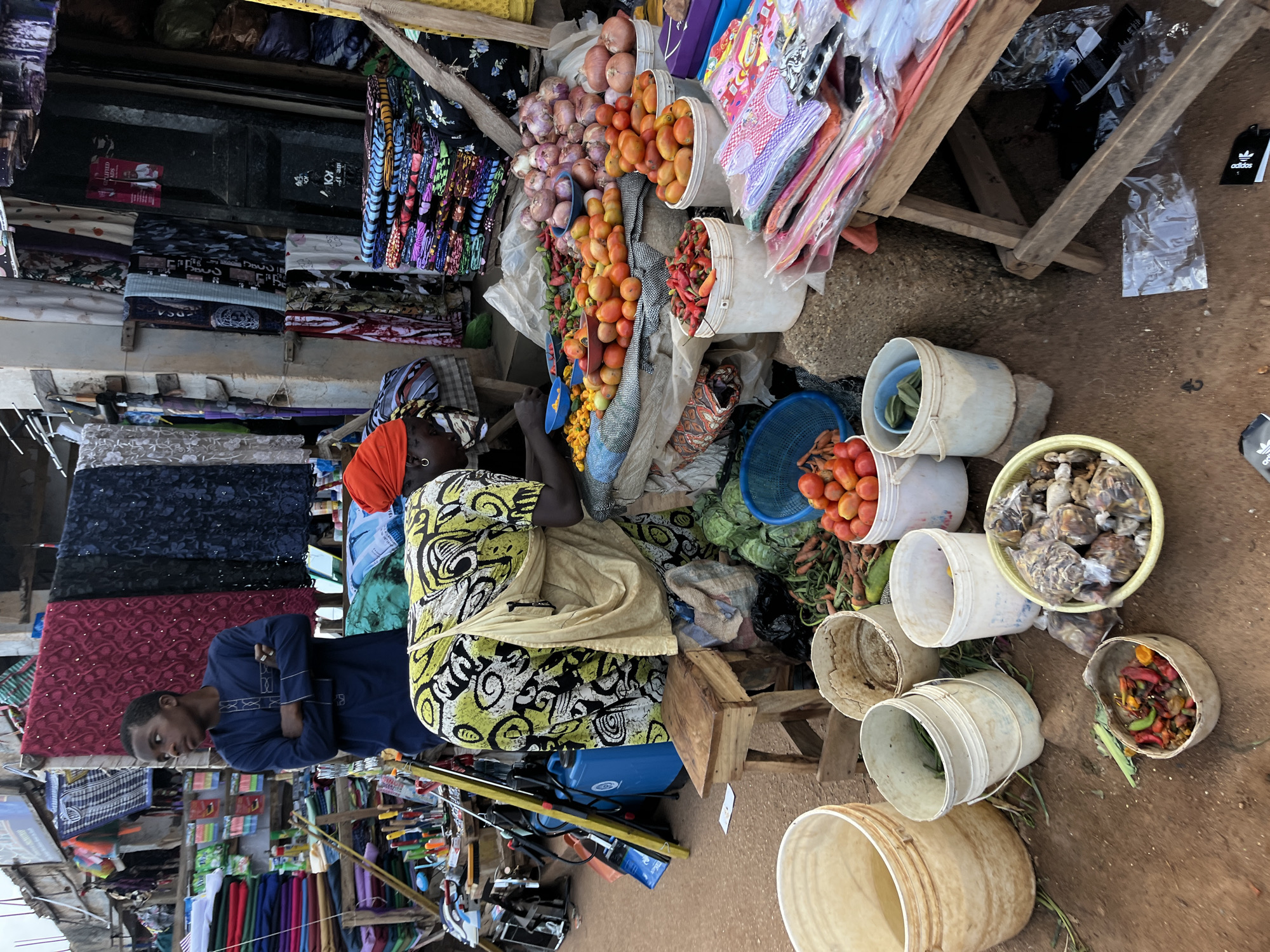 A busy market scene is depicted in Bukuru, Nigeria, with an outdoor stall featuring a variety of goods for sale. In the foreground, several large, white plastic buckets are placed on the dusty ground, some appearing to be empty or partially filled with what might be water or produce. To the left, a young boy wearing a dark blue, traditional African tunic stands with his arms crossed, looking to his left with a neutral expression.
Dominating the center of the image is a woman, partially obscured by her clothing, wearing a bright orange headwrap and a patterned dress with yellow, green, and black designs. She is tending to a display of fresh produce. Piles of red tomatoes, green peppers, onions, and other vegetables are arranged on a textured surface, possibly a tarp or cloth, in front of and beside her. Several white buckets are filled with these vegetables.
Behind the produce stall, the market continues with more stalls and wares. Colorful fabrics, possibly for clothing, are displayed on racks and hung overhead. Stacks of folded textiles in various vibrant patterns are visible, suggesting a textile market. Various tools, including what appear to be knives and possibly a spray bottle, are also present in the background, indicating a diverse range of goods being sold. The ground is a mix of dirt and scattered debris, typical of an outdoor market environment. The overall atmosphere is one of commerce and community activity.