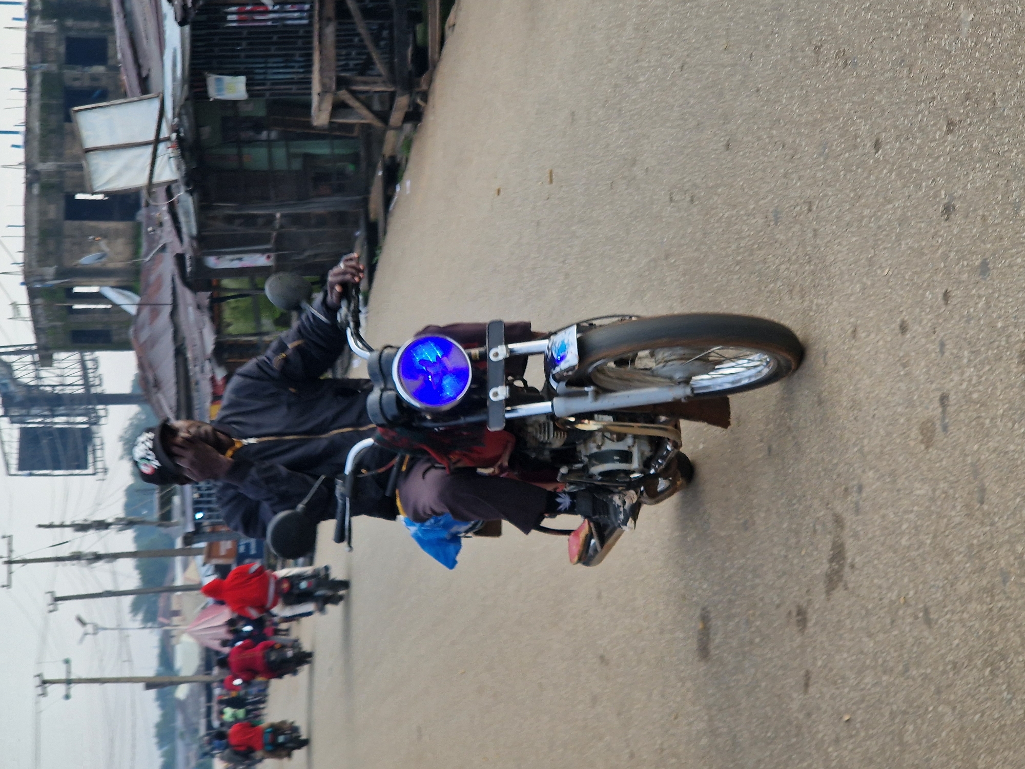 The image depicts a scene in Kabong, Nigeria, likely a roadside environment. A man is riding a motorcycle in the foreground, his right hand on the handlebars. He is wearing a dark jacket, dark pants, and a black baseball cap with a design on the front. The motorcycle has a bright blue headlight. In the background, several other individuals are riding motorcycles, many of them wearing red hooded jackets. The environment shows signs of urban development with buildings and structures visible, some appearing unfinished or dilapidated. Power lines and poles are also present, indicating infrastructure. The road surface is asphalt, with some scattered debris. The overall impression is a busy street scene in a developing urban area.