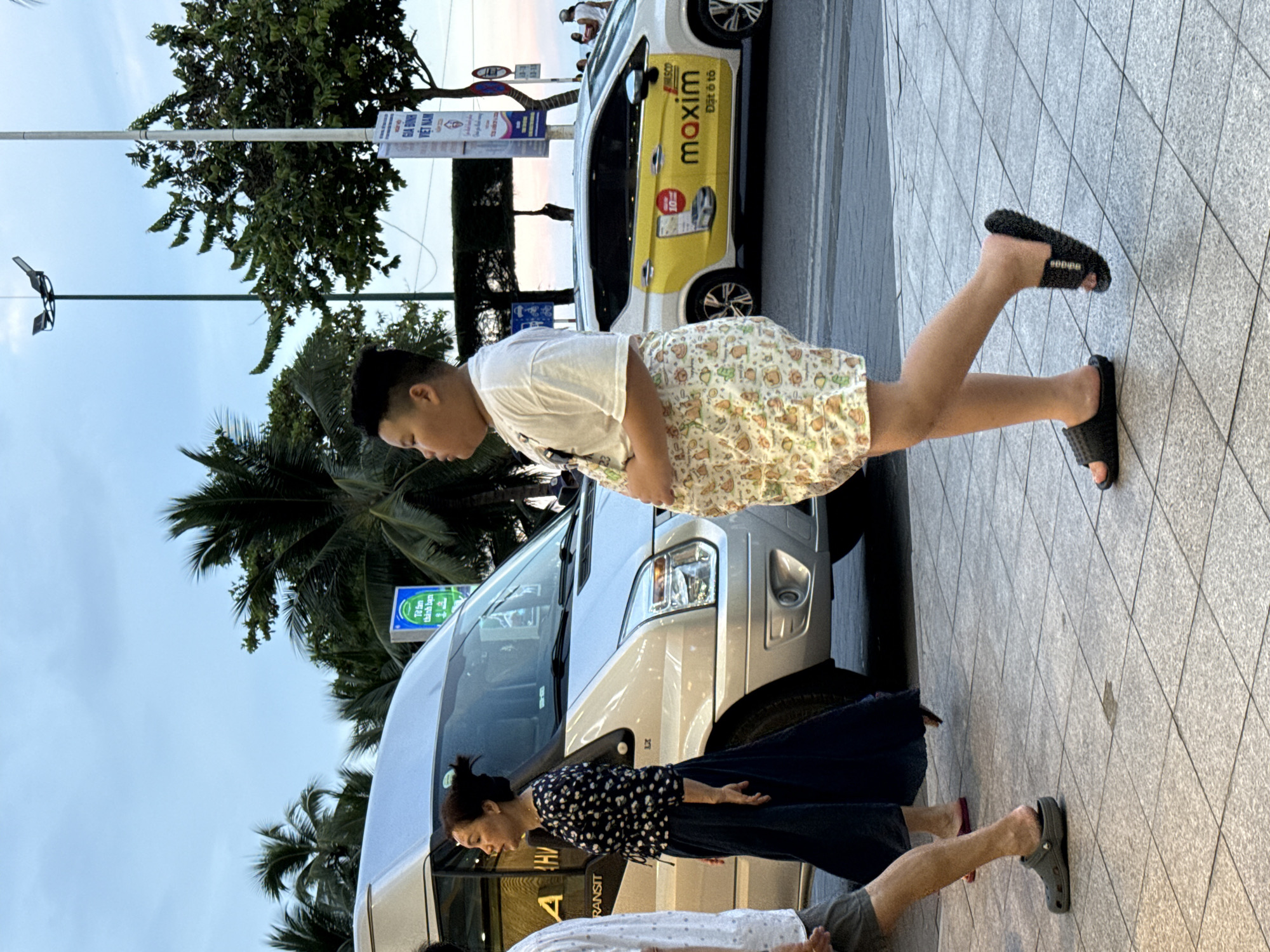 The image shows a group of people on a sidewalk in Nha Trang Ward, Vietnam. A young boy in a white t-shirt and patterned shorts is standing with his legs spread apart, as if in mid-movement. Next to him, a woman in a dark, floral-patterned dress and a man in gray shorts and a white t-shirt are also on the sidewalk. Behind them, a silver van is parked, and further back, a yellow car with the Maxim logo is visible. Palm trees line the street, suggesting a tropical or subtropical location. The sky is overcast, indicating it might be early morning or late afternoon. The scene appears to be a casual street environment, possibly a tourist area given the presence of taxis and the general ambiance. The people's postures suggest they are either walking, posing for a photo, or engaged in some spontaneous activity. The Maxim car has text in yellow and black. The van has "TRANSIT" written on its side. There are also signs with Vietnamese text, including one with "VIET NAM."