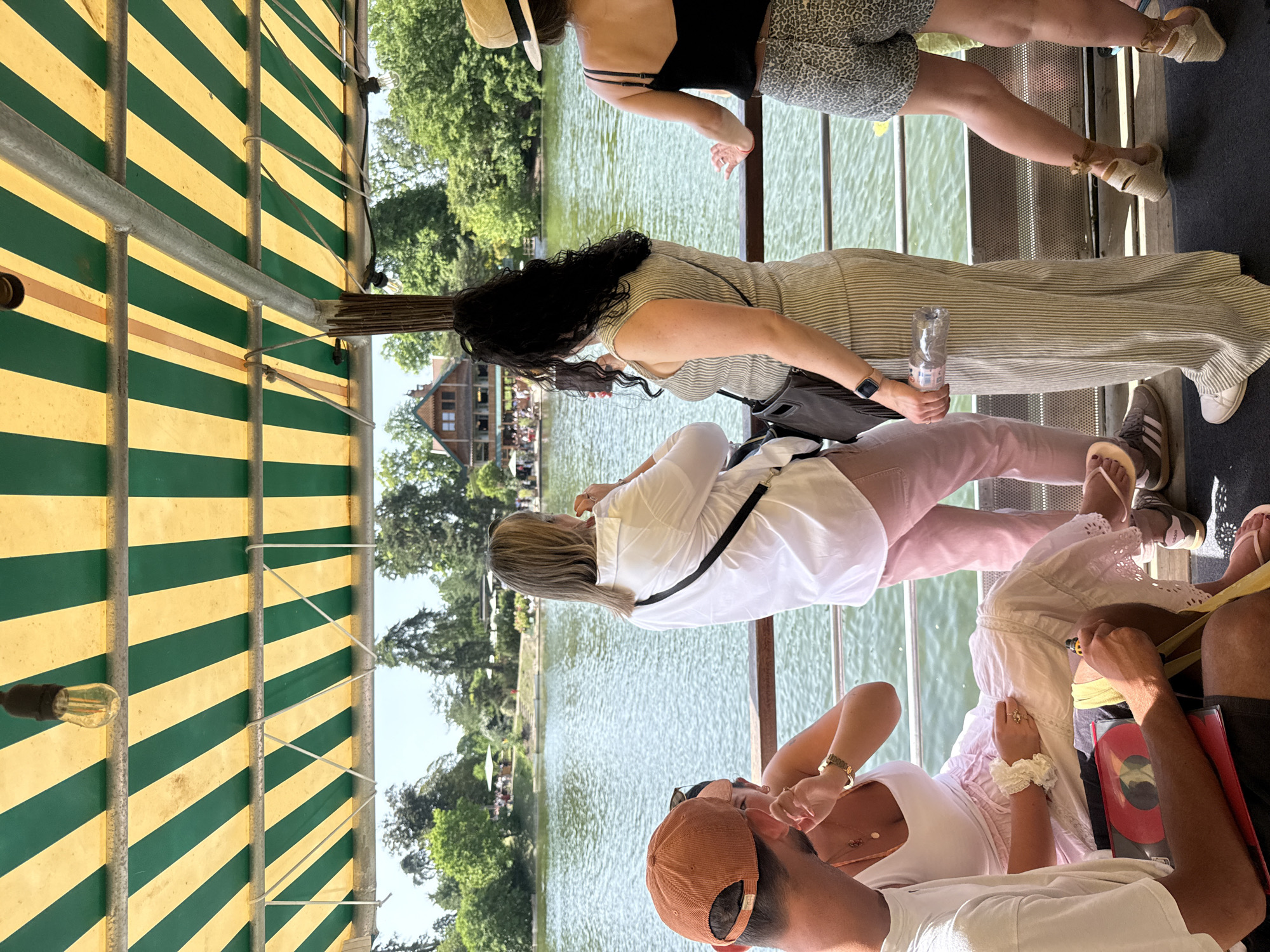 The image shows a group of people on a boat on a body of water. A building and trees are visible in the background. The boat has a striped yellow and green awning. The scene is in Paris, France.