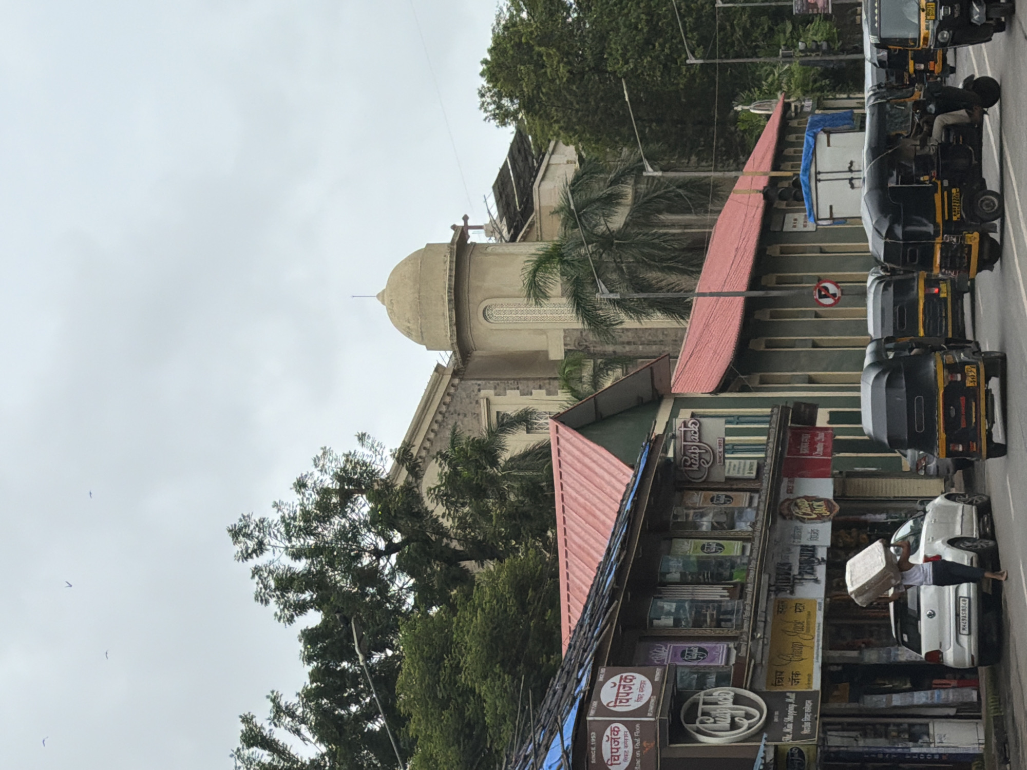 The image captures a street scene in Mumbai, India, likely on a cloudy day. In the foreground, several auto-rickshaws are present, with one in the center driven by a person. To the left, a white Volkswagen Polo car is parked, and a person is walking away from it carrying a large, rectangular box on their head. Adjacent to the car, a row of shops is visible with various signs, including "Cheap Jack" and "The Mini Shopping Mall". The shop awnings appear to be made of corrugated metal and blue tarpaulin.
In the background, a large building with a distinct dome and a cross is prominent, suggesting it is a religious structure like a church or cathedral. The architecture is colonial-era. Trees, including palm trees, are scattered throughout the scene. The sky is overcast with a few birds visible in the upper portion of the image. The overall impression is of a bustling urban environment with a mix of commercial establishments and a significant religious landmark.