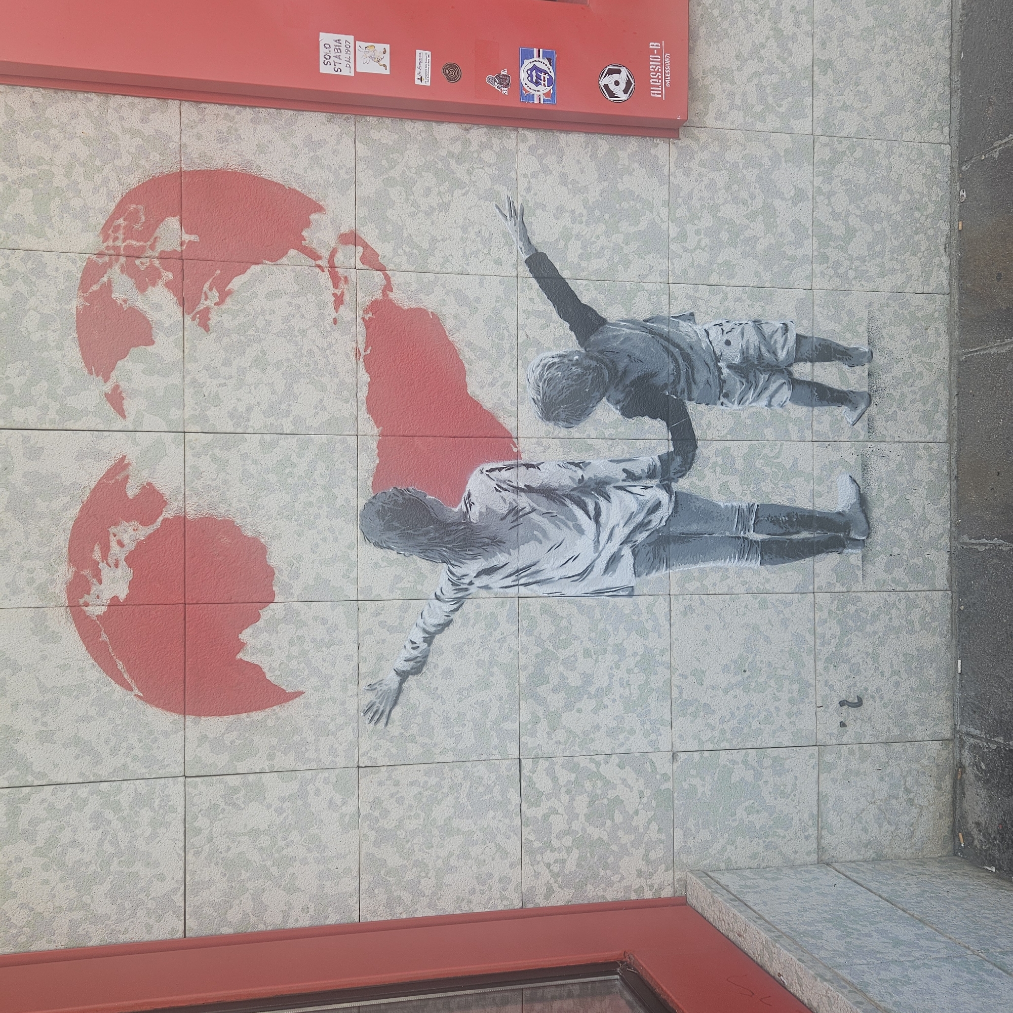 The image shows a mural painted on a tiled wall. The mural depicts two children, a girl and a boy, holding hands and reaching towards a red heart-shaped globe. The girl is on the left, wearing a long-sleeved shirt and pants, and the boy is on the right, wearing a t-shirt and shorts. The globe appears to be stylized, with continents outlined in white. To the right of the mural, there is a red vertical panel with several stickers on it. One sticker reads "SOLO STABIA DAL1907". Another sticker has a logo that resembles a soccer ball with wings. The bottom of the mural shows the names "ALESSIO-B" and "@AlessioB71". The mural is located on a street in Bastia, Italy, specifically near the ferry terminal. The ground in front of the mural is paved and wet, with some cigarette butts visible. The wall itself is made of light-colored tiles with a speckled pattern.