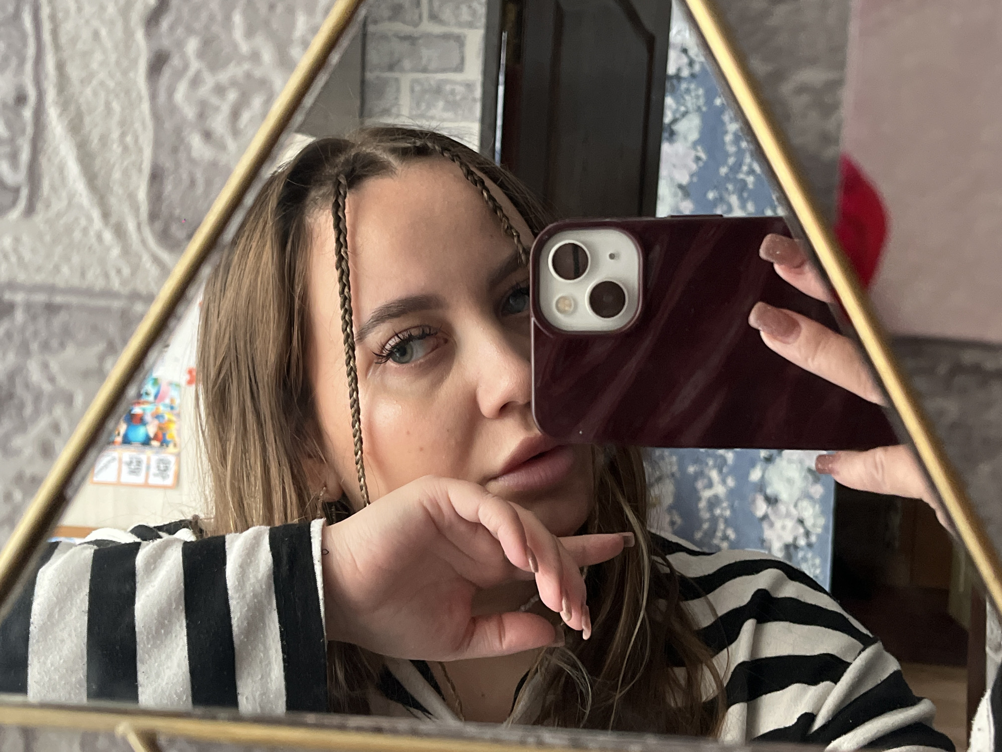 A young woman with long brown hair and braids is taking a selfie in a mirror with a triangular frame. She's holding a dark red phone with a dual-lens camera. She wears a black and white striped shirt and has natural-colored nails. The background shows a glimpse of a brick wall and what appears to be a patterned wall hanging. The photo seems to be taken indoors, likely in a private residence. There is no indication that the photo was taken in Novopokrovskaya, Russia.
