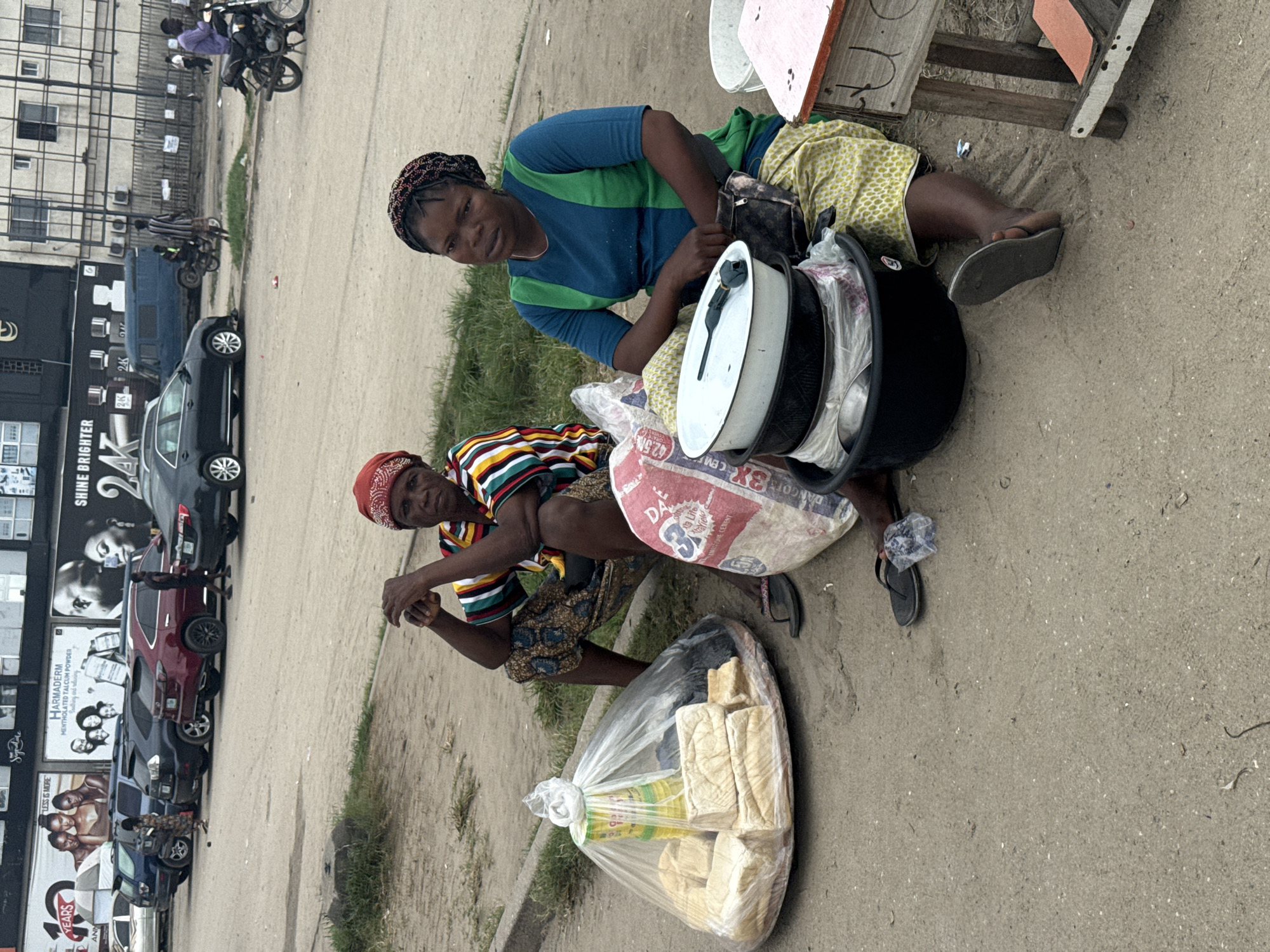 In Satellite Town, Nigeria, two women are seated on the ground next to their merchandise. The woman on the left, wearing a red headscarf and a striped shirt, sits with her hands clasped. In front of her is a large plastic bag filled with loaves of bread and some yellow containers. To her right, a woman in a blue and green top and a patterned skirt is seated, looking forward. She has a black pouch around her waist. In front of her are several stacked bowls, a black basin, and a bag of what appears to be cement. A small wooden stand with a pink surface is to her right. The ground is dirt and sand, with a curb and a paved road visible in the background. Several cars are parked on the road, and a few people are walking or riding motorcycles in the distance.