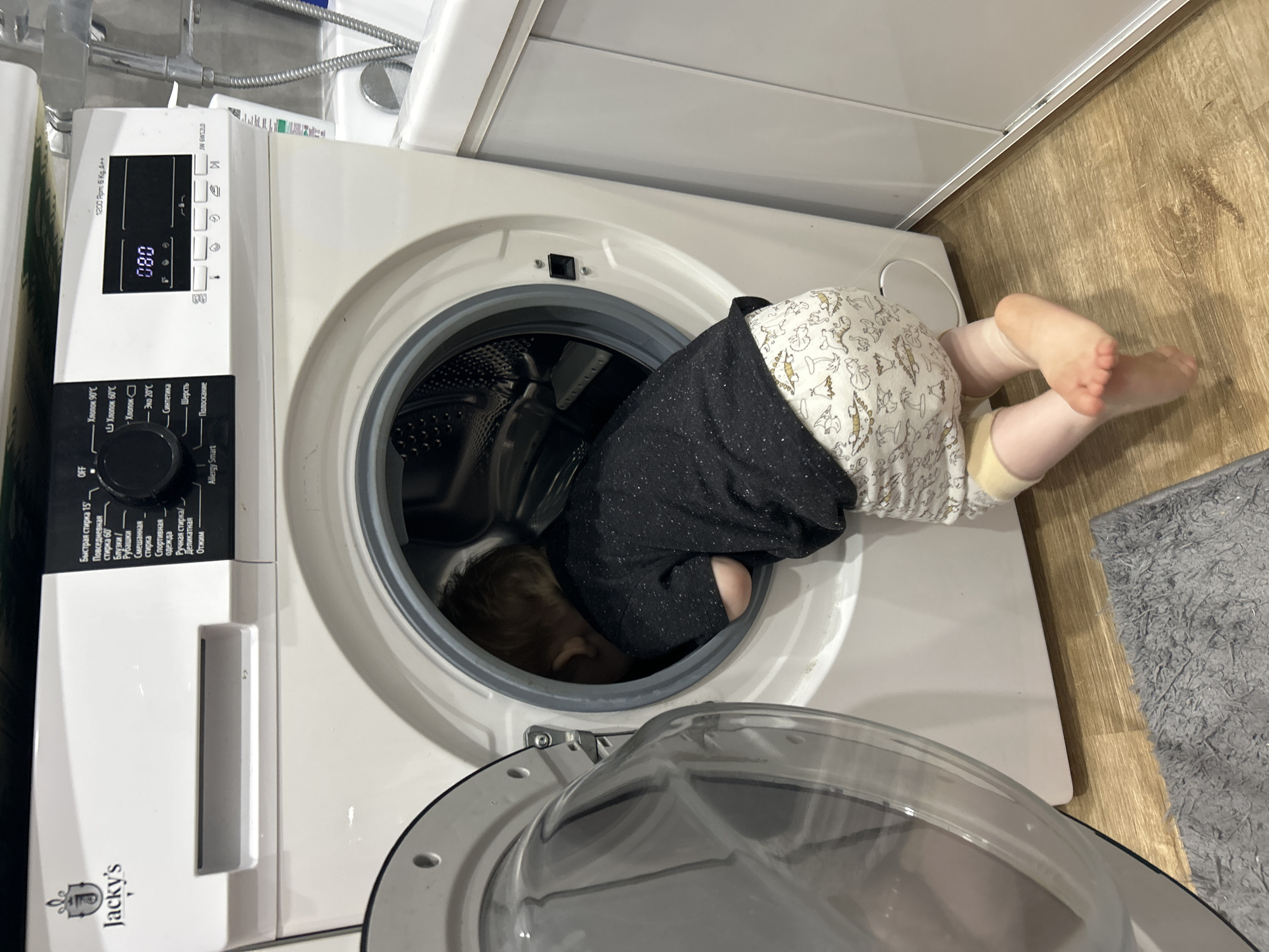 A curious young child is depicted in a domestic setting, with their upper body playfully tucked inside the open drum of a front-loading washing machine. The child is wearing a dark, possibly sparkly, top and light-colored shorts or pants adorned with cartoon-like patterns. Only their legs, bare feet, and patterned lower attire are visible outside the appliance, suggesting they are fully exploring the interior of the machine.
The washing machine itself is white, with a clear circular door that is currently open. Its control panel is visible, featuring a digital display showing "080" and a prominent dial with various wash cycle options written in Russian. Visible text includes the brand "Jacky's" and model specifications like "1200 Rpm. 6 Kg. A++" and "JW 6W1ZLD." Cycle options on the dial include "Быстрая стирка 15'" (Quick wash 15'), "Повседневная стирка 60°" (Daily wash 60°), "Блузки / Рубашки" (Blouses / Shirts), "Смешанная стирка" (Mixed wash), "Спортивная одежда" (Sportswear), "Ручная стирка / Деликатная" (Hand wash / Delicate), "Отжим" (Spin), "OFF", "Allergy Smart", "Хлопок 90°С" (Cotton 90°C), "Хлопок 60°С" (Cotton 60°C), "Хлопок" (Cotton), "Эко 20°С" (Eco 20°C), "Синтетика" (Synthetics), "Шерсть" (Wool), and "Полоскание" (Rinse).
The setting appears to be a laundry area or bathroom within a home in Chaikovsky, Russia. The floor is light-toned wood, possibly laminate, and a grey, shaggy rug is partially visible on the right side. Behind and above the washing machine, a white structure, likely part of a bathtub or sink unit, is visible, along with some chrome plumbing fixtures. The lighting suggests an indoor, daytime scene. The image captures a moment of childhood curiosity and exploration.