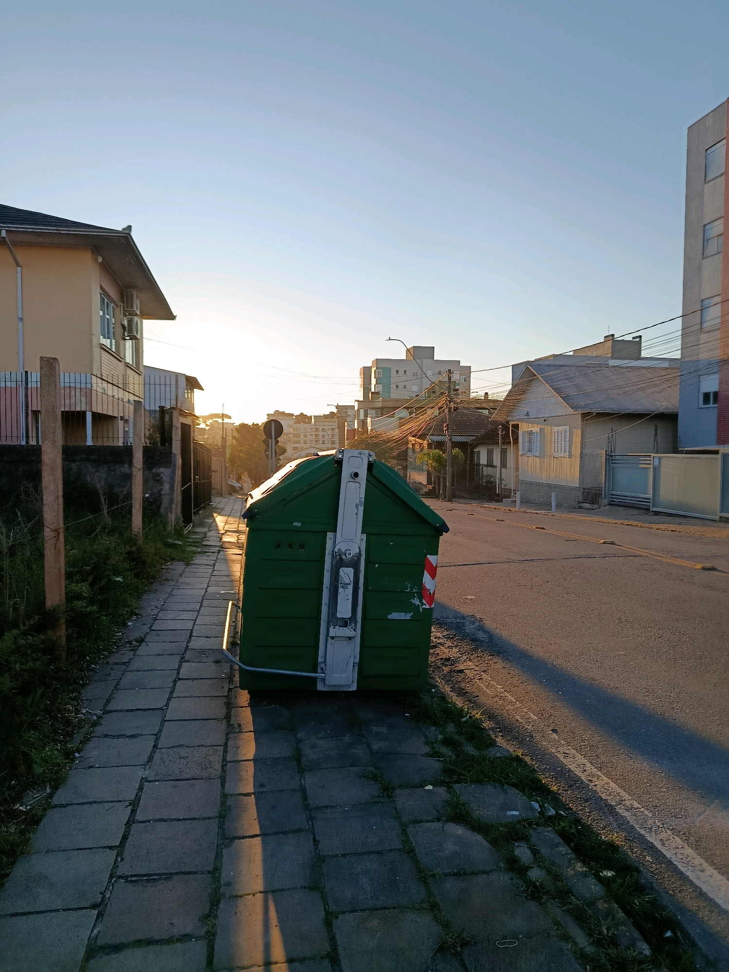 The image depicts a street scene at sunrise in the city of Caxias Do Sul, Brazil. A large, green dumpster is positioned on a paved sidewalk next to a street. The dumpster has white and red reflective markings on its side. The sidewalk is made of square concrete pavers. To the left of the sidewalk is a grassy area with some vegetation and a wooden fence. Behind the fence is a two-story building with a light-colored facade, several windows, and air conditioning units visible. Across the street, there are residential buildings and a multi-story building in the distance. The sky is clear and blue, with the sun low on the horizon, casting long shadows across the scene. Overhead, a network of electrical wires is visible, strung between utility poles. The environment appears to be an urban or suburban residential area.
