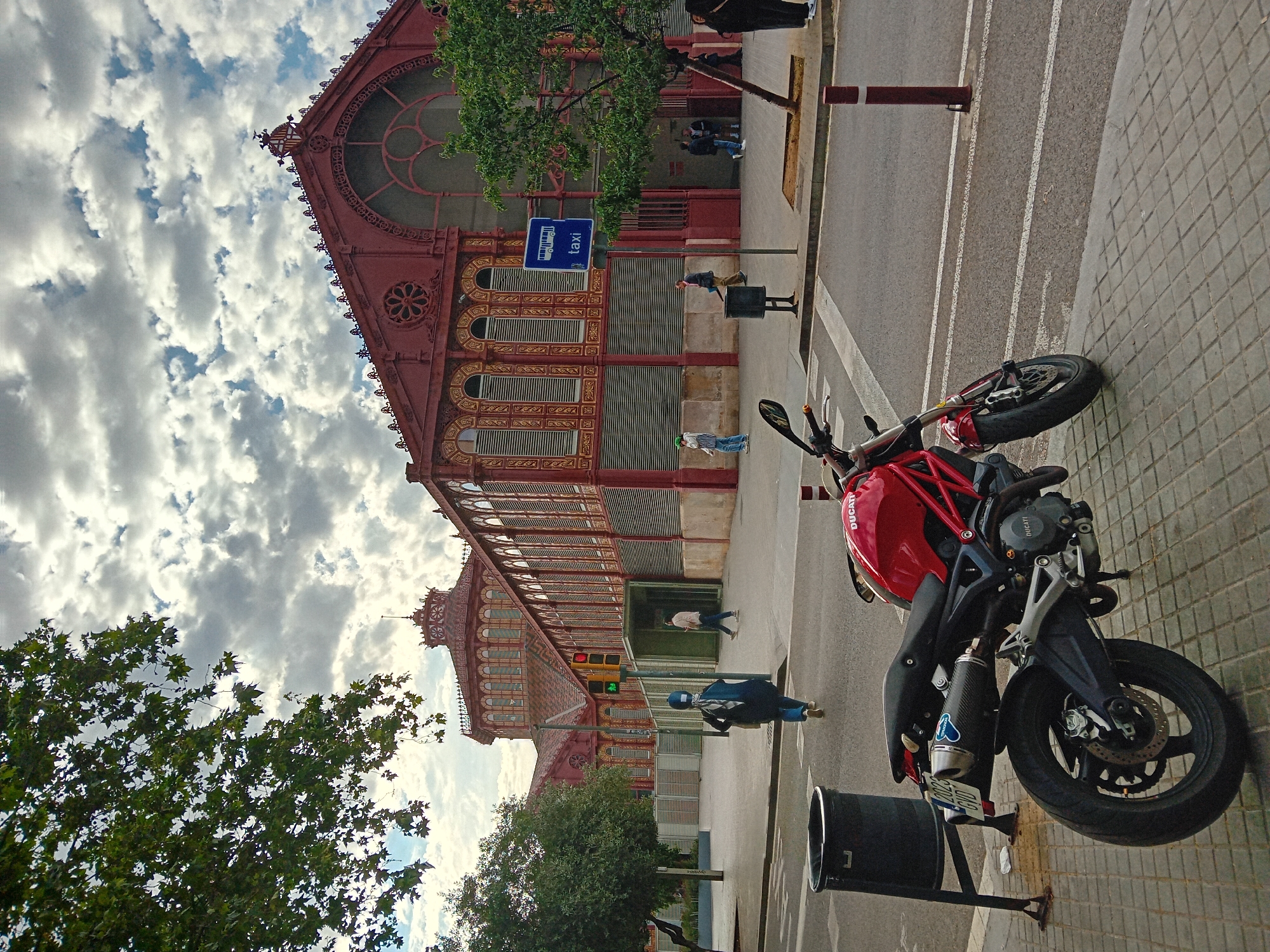 The image depicts a street scene in Barcelona, Spain, featuring the El Born Cultural and Memorial Center. This distinctive brick building is visible on the left, its ornate windows and architectural details in clear view against a cloudy sky. Parked nearby on the right is a red Ducati motorcycle, its front wheel close to a trash can. Pedestrians walk along the sidewalk in the background. A street with painted white lines is between the sidewalk and building.