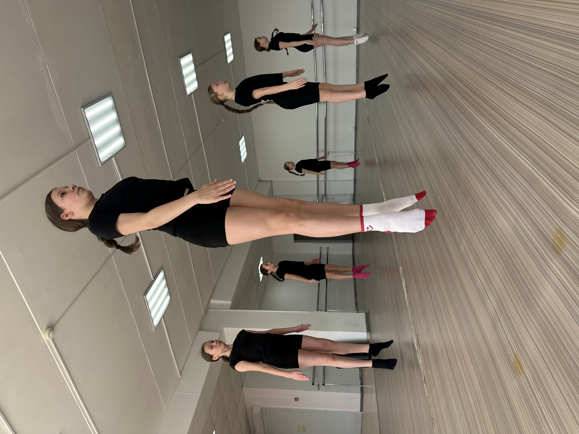 The image depicts a dance studio in what appears to be Votkinsk, Russia. Five young women are practicing a routine. They are wearing black shorts and tops and varying socks. The studio is mirrored, reflecting the dancers. The floor is a light brown wood grain, while the walls and ceiling are white. There are rectangular ceiling lights. The photo captures a moment of stillness in the dance practice.