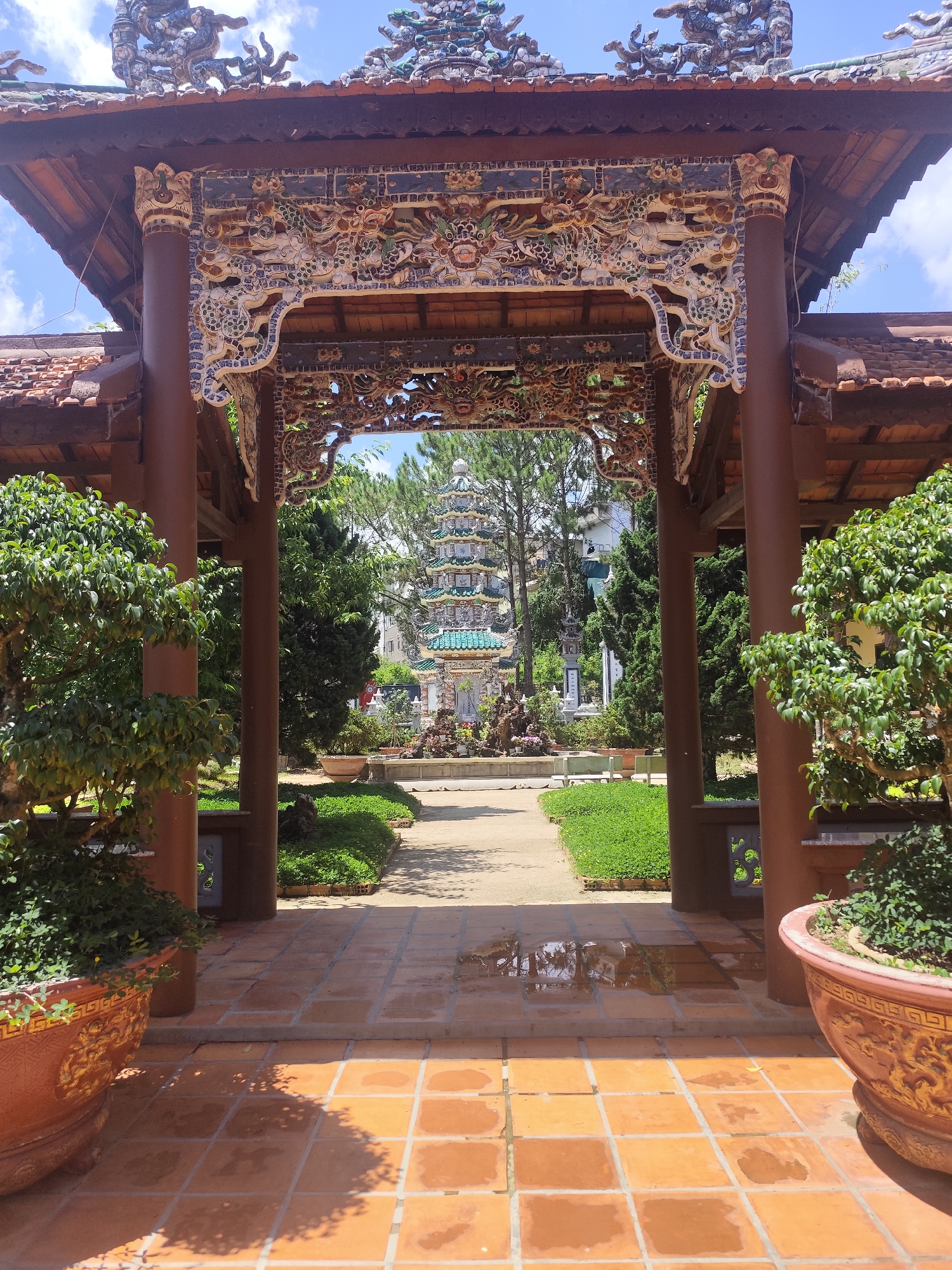 The image shows a tiled patio area under an ornate wooden structure, framing a view of a pagoda in the city of Da Lat, Vietnam. The structure features red wooden posts and beams, adorned with intricate carvings and colorful mosaic detailing. The pagoda is visible in the distance, a multi-tiered structure with green and blue tiled roofs. The patio is covered in square orange-red tiles, some of which have puddles of water. Large potted bonsai trees are positioned on either side of the patio, framing the view. Beyond the patio, a pathway leads towards the pagoda, flanked by manicured green lawns and various trees and shrubbery. The sky above is blue with scattered white clouds.