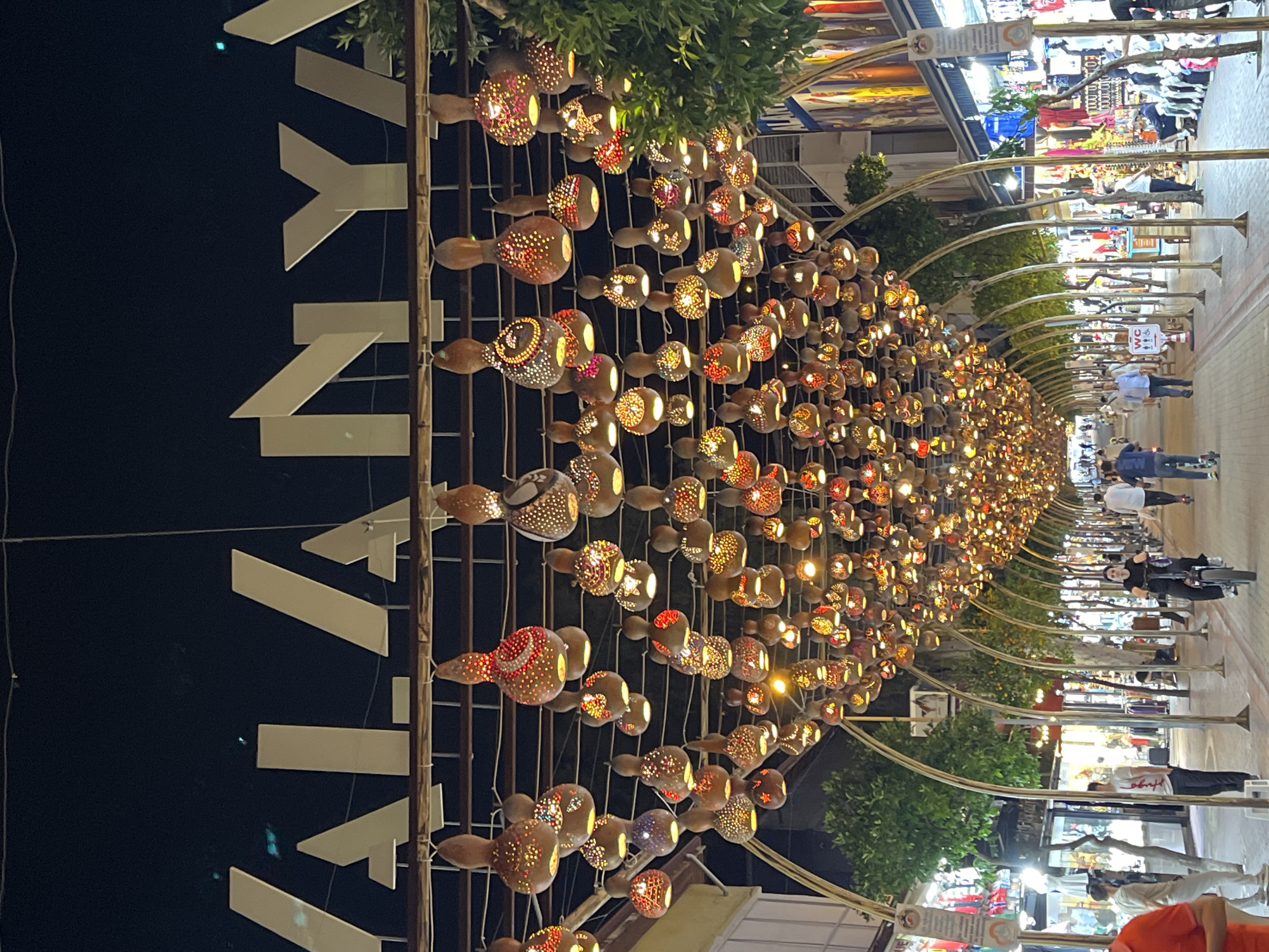The image captures a vibrant pedestrian street or market in Alanya, Turkey, at night. The scene is dominated by an elaborate overhead lighting installation consisting of numerous ornate, gourd-like lamps. These lamps are intricately patterned with holes, allowing warm, colorful light to emanate, creating a tunnel-like effect along the thoroughfare. They are strung along metal archways that span the width of the street.
On the far left, large, white, angular letters are mounted vertically, partially spelling out "Y" and "N," which are part of the word "ALANYA," indicating the location. Green foliage, likely from trees or potted plants, is visible along the side, interspersed with the metal arches and lights, adding a natural element to the urban setting.
Below the illuminated canopy, the street is bustling with several people. Individuals are seen walking and standing, some appearing to be browsing the various goods displayed in the shopfronts that line both sides of the street. One person is visible on a scooter, suggesting movement within the area. The shops showcase a variety of items, though specific details are indistinct. Further down the street, a "WC" sign is visible on a pole. The overall impression is that of a lively evening promenade or shopping area with a distinctly festive and welcoming ambiance.