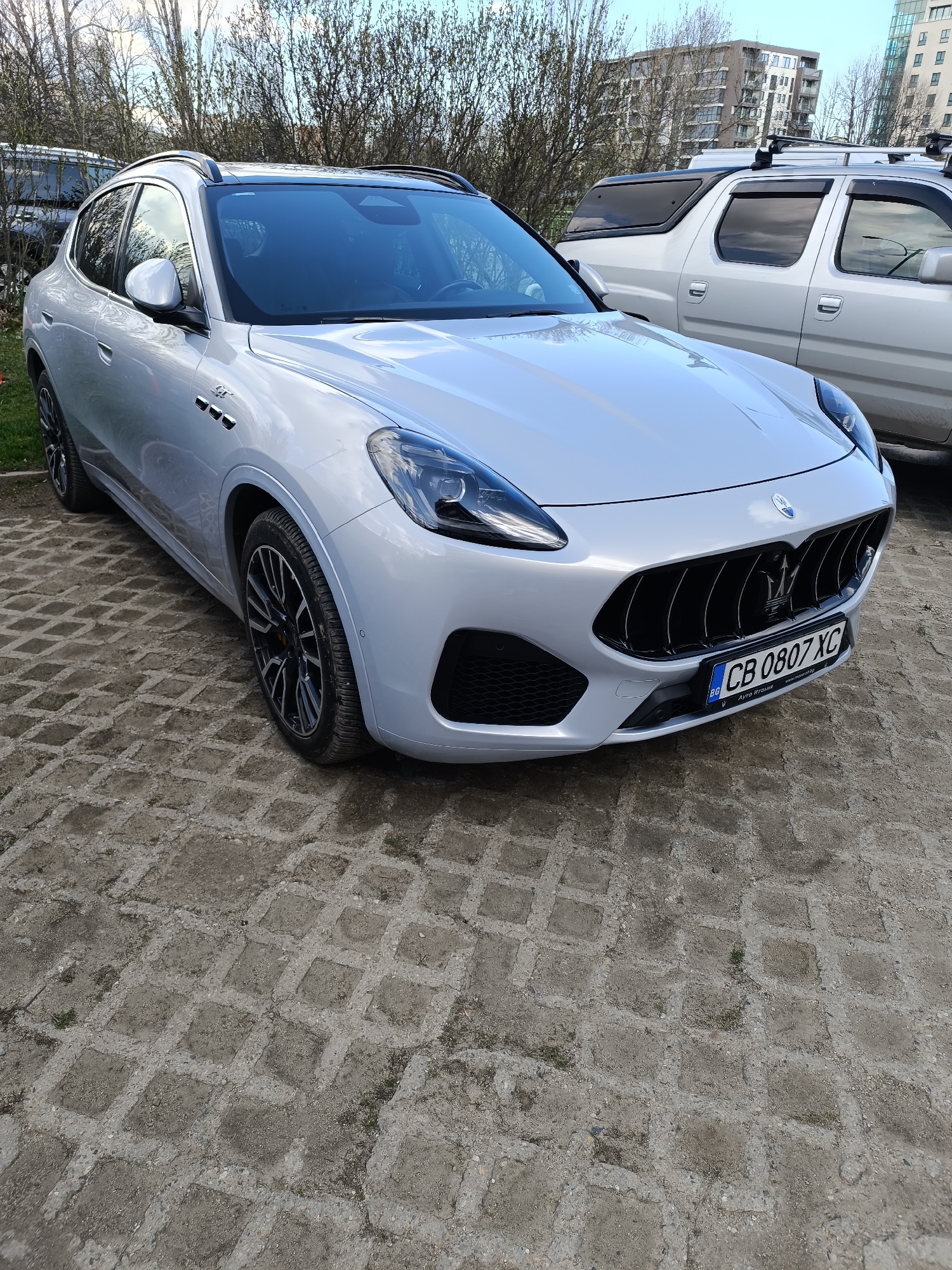 The image shows a silver Maserati Grecale SUV parked on a paved area. The SUV is the main subject, displaying its front design with a prominent Maserati grille and license plate reading "CB 0807 XC." The vehicle's wheels have a black and silver spoke pattern. In the background, there are trees with bare branches and an SUV with a roof rack, suggesting an outdoor setting. Apartment buildings are also visible in the distance. The environment suggests an urban or suburban area. The image focuses on the parked vehicle and its immediate surroundings.