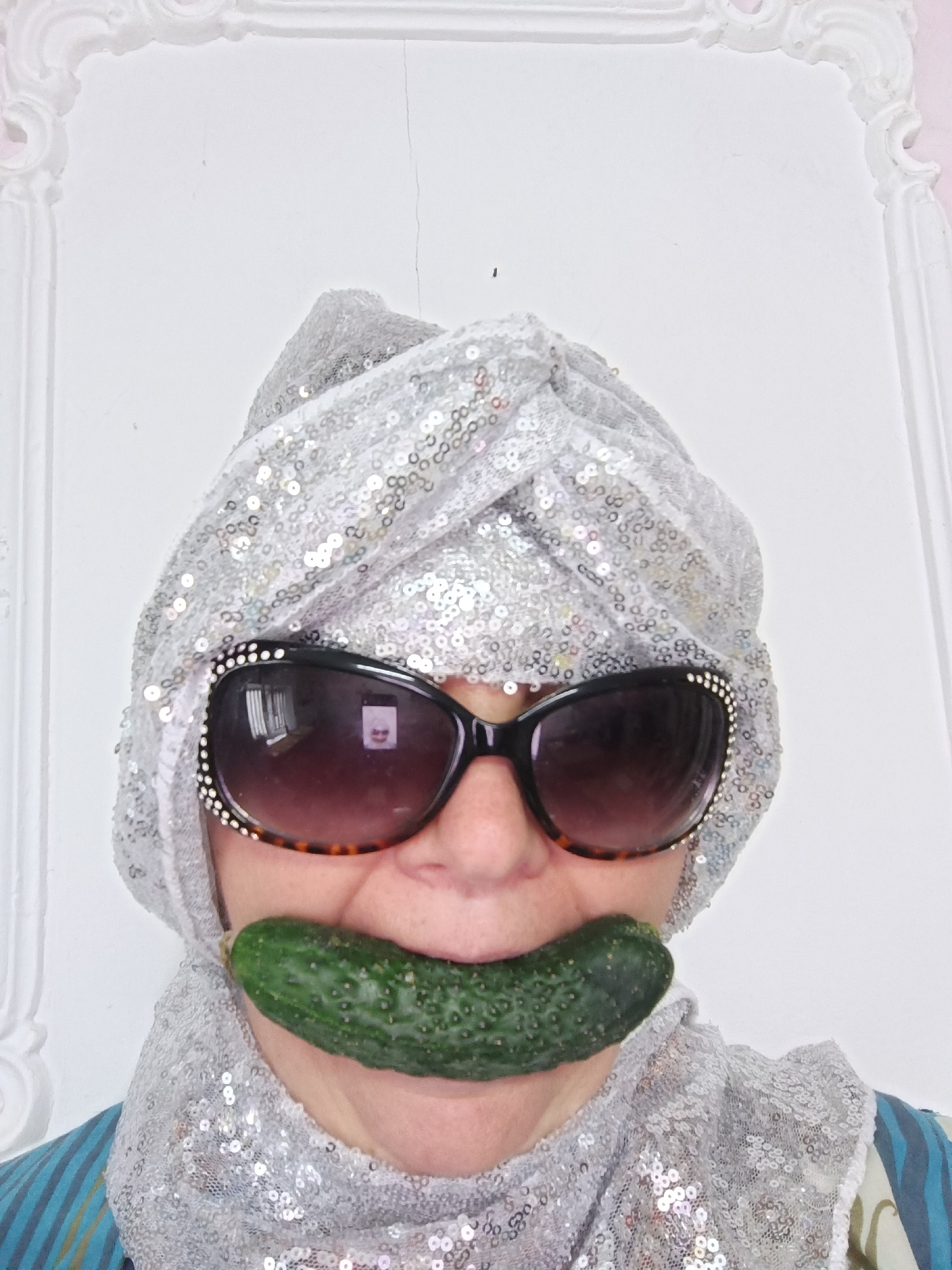 In Tyshky, Ukraine, a person wearing a silver sequined turban and scarf, and large sunglasses with rhinestones, has a pickle placed horizontally across their mouth. The person is facing forward, and the background is a white wall with decorative molding. The image is a medium shot, focusing on the person's face and upper body.