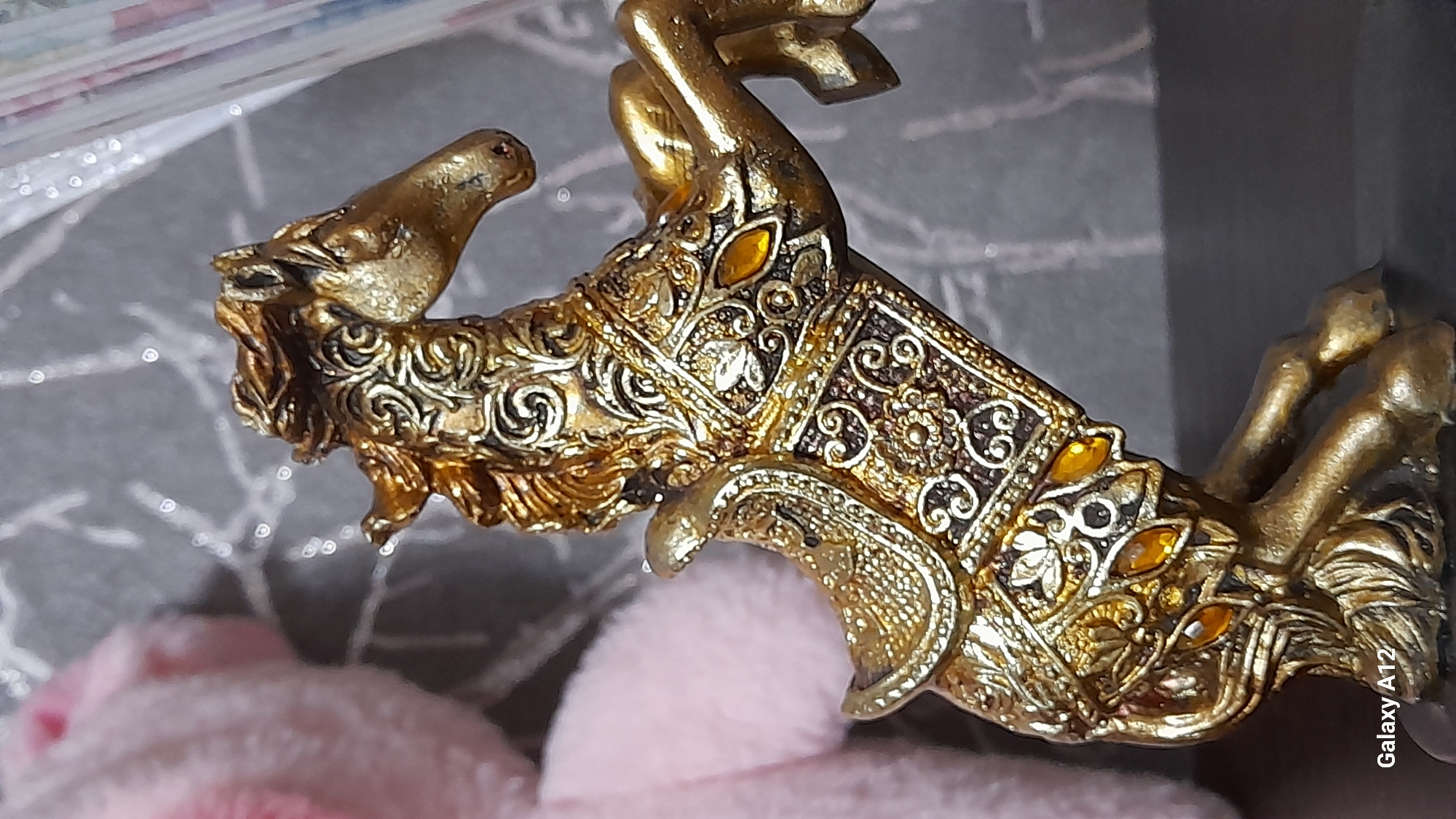 The image displays an ornate golden statuette of a rearing horse, captured in a close-up, vertical perspective. The statuette features intricate, filigree-like patterns with floral and scrolling motifs covering its entire body, including the mane, tail, and decorative barding. Multiple small, oval, amber-colored gem-like accents are embedded within these patterns on the horse's side and legs. The horse is depicted in a dynamic pose, with its front legs lifted and its hind legs planted on a dark, flat base. This base rests upon another dark, unidentifiable surface.
In the background, to the immediate left of the statuette, a soft, out-of-focus pink object, possibly fabric or a plush item, is partially visible. The central and right background consists of a blurred, mottled grey surface, exhibiting diffuse white specks and bokeh effects. A vertical, semi-transparent, blurred object extends along the far right edge of the frame. The text "Galaxy A12" is inscribed in white in the bottom-left corner of the image, identifying the camera used. This scene is located in the city of Марковский, Russia.