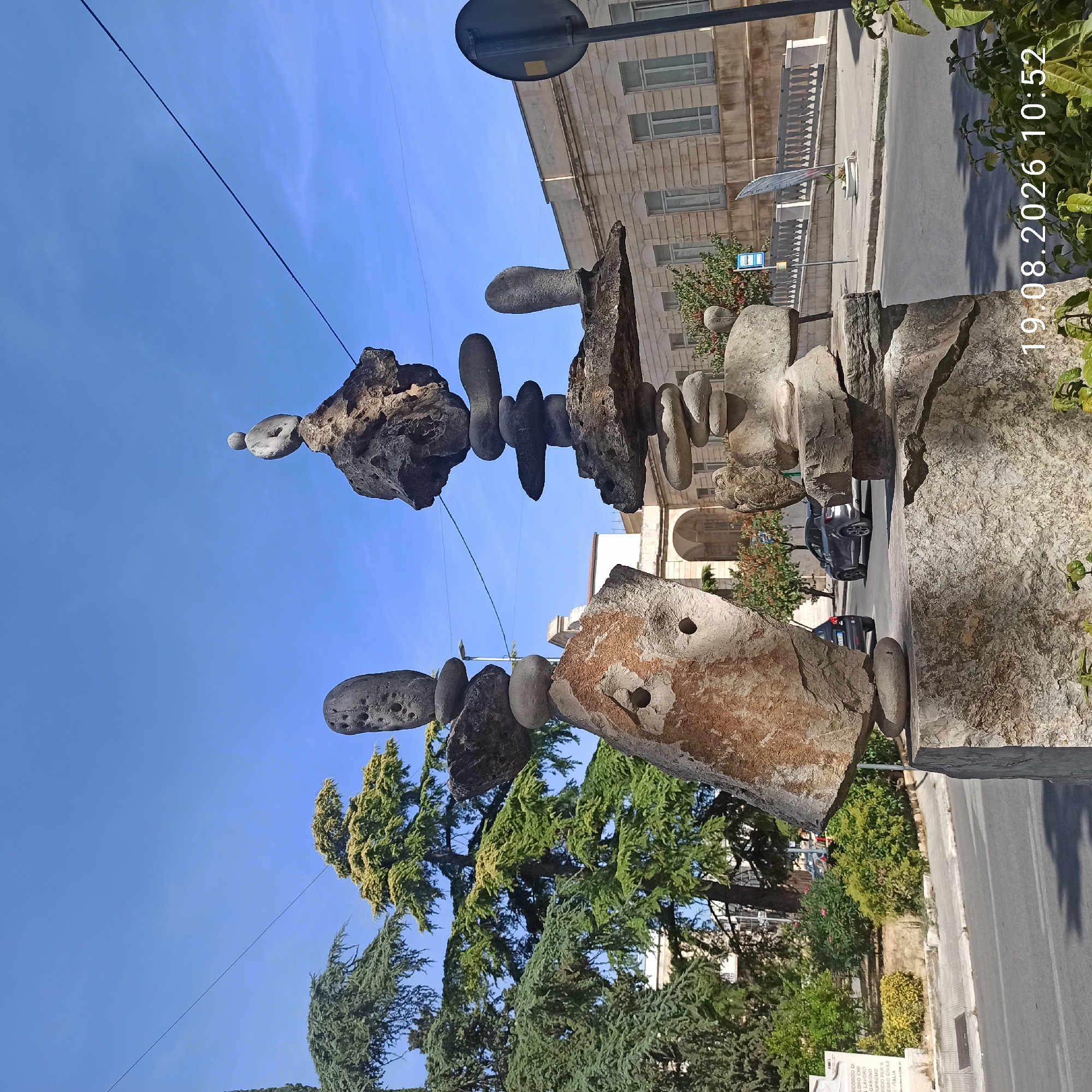 The image shows a contemporary stone sculpture in the city of Ruvo di Puglia, Italy. The sculpture consists of several large, irregularly shaped stones precariously balanced on top of each other, creating abstract figures. One large stone in the foreground on the left has two circular indentations that resemble eyes, giving it a face-like appearance. To the right, another, taller structure of balanced stones is topped by a single round stone with a hole through its center.
The sculpture is situated outdoors, likely in a public space, with a paved road and sidewalk visible. In the background, there are trees with green foliage, a light-colored building with windows, and a street sign. Two cars are visible on the road. The sky is clear and blue. A watermark in the lower right corner reads "19.08.2026 10:52".