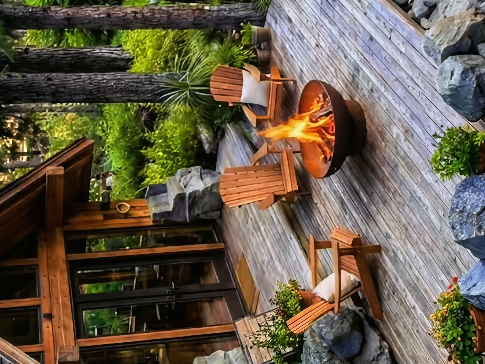 This rotated image, captured in Sofiivska Borshchahivka, Ukraine, depicts a cozy outdoor living space on a wooden deck adjacent to a rustic wooden house or cabin, likely in the evening or at night. The scene is set amidst abundant green foliage and tall tree trunks, suggesting a natural, forested environment.
In the center of the wooden deck, a dark metal fire pit holds a strong, blazing fire, casting a warm orange glow. Several wooden Adirondack-style chairs with white cushions are arranged around the fire pit, inviting relaxation. Two chairs are clearly visible in the foreground and midground, positioned to face the fire. The deck itself is constructed from light-colored wooden planks, weathered and textured.
To the left, a section of a wooden house or cabin is visible, featuring large windows framed by dark wood, reflecting some of the surrounding greenery. The architecture harmonizes with the natural setting. Potted plants with vibrant green foliage and small red or yellow flowers are strategically placed on the deck and around the rocky landscaping that borders the area. Large, grey boulders and smaller rocks are scattered along the edges of the deck, integrating natural elements into the design. Tall, dark tree trunks rise vertically in the background, surrounded by lush green undergrowth and moss.
The overall atmosphere conveyed is one of warmth, tranquility, and natural beauty, perfect for an evening gathering or quiet contemplation by the fire. No people are visible in the image, and there is no discernible text.