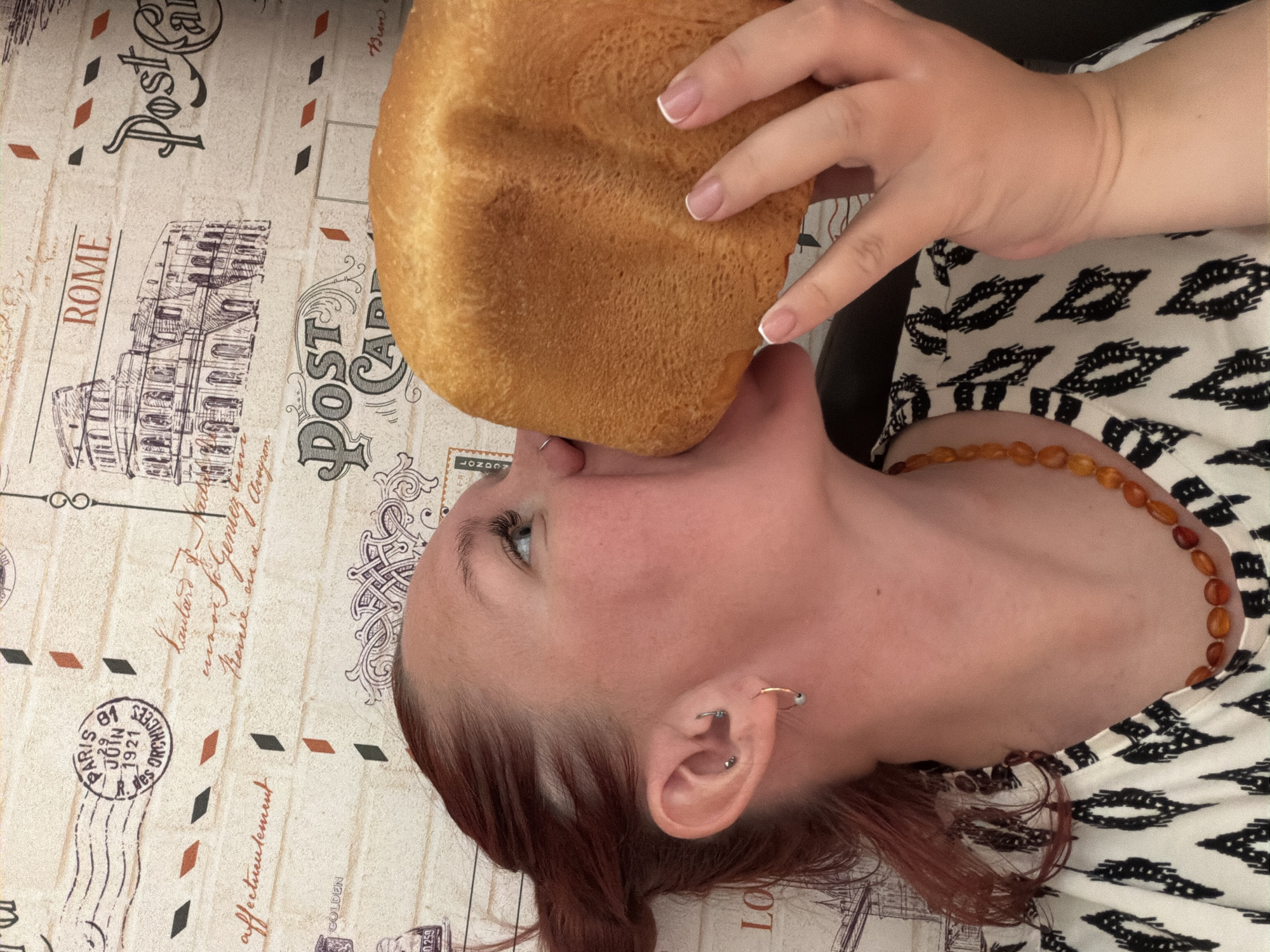 A person is positioned in front of a patterned surface featuring postcard imagery, holding a loaf of bread with their hand. The person appears to be bringing the loaf toward their mouth. The exact location of the scene in the city of Chaikovsky, Russia, is not discernible from the image.