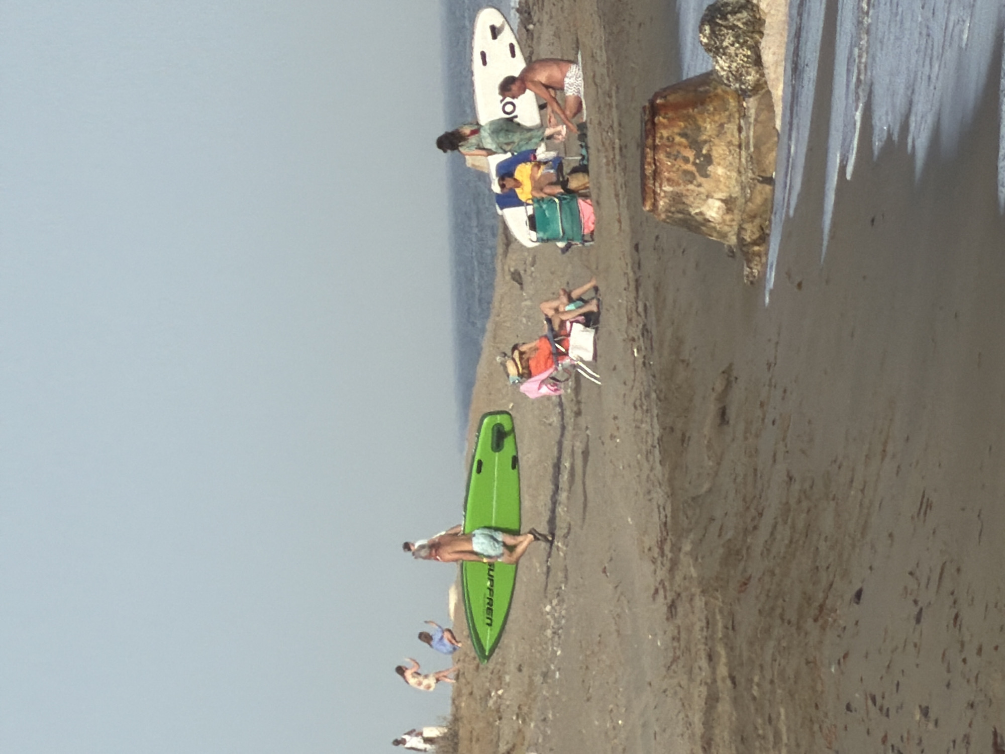 A group of people are on a sandy beach in El Velerín, Spain. The sky is clear and pale blue, with a hint of haze. The sea is a muted blue-grey and appears calm. In the foreground, waves gently lap the shore. Several individuals are scattered across the sand.
To the left, a man in blue swim trunks carries a bright green surfboard. Behind him, a woman in a patterned dress and another in a blue top are crouched down. Further left, a person in a white garment stands near some foliage.
In the middle of the frame, several people are gathered. A woman in an orange sunhat sits in a beach chair. Next to her, another person in a yellow shirt is seated in a blue and green beach chair, with a large green bag next to them. A woman in a patterned green dress stands near a white surfboard, and a man in white patterned swim trunks sits on the sand to the right of the surfboard.
Further to the right, a white surfboard with black accents is propped up. In the far right of the image, a man in white patterned swim trunks is seated on the sand.
In the mid-ground, a large, weathered, conical concrete structure with orange and brown staining sits partially in the sand and partially in the shallow water. To its right, a large, greyish rock is visible. The sand itself is a light brown and appears somewhat coarse. Scattered on the sand are small pieces of debris and seaweed.