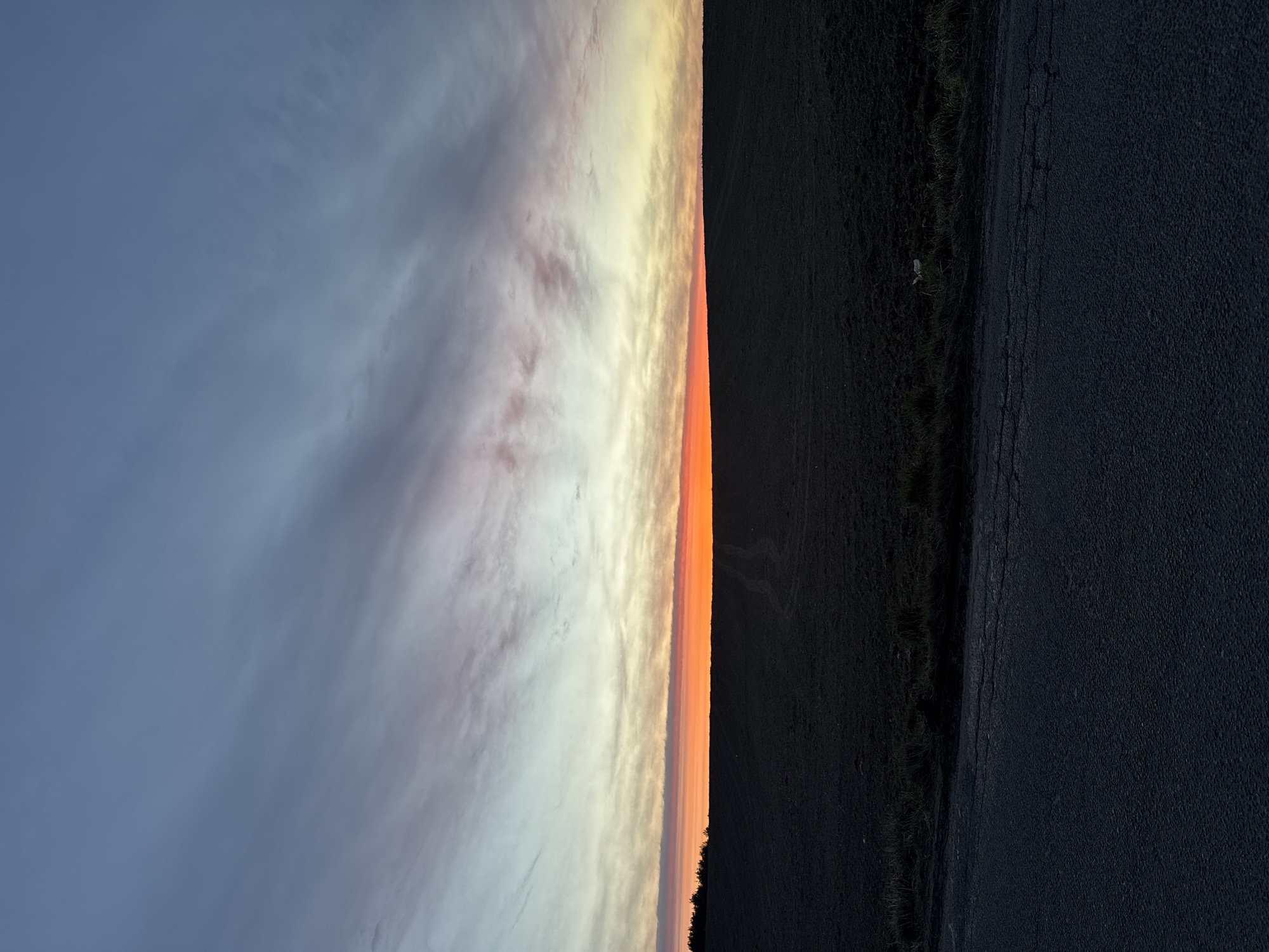 A landscape photograph taken in Ждановіцкі сельскі Савет, Belarus, shows a horizon at sunset. The left side of the image depicts a gradient of blue sky fading into wispy clouds tinged with pastel shades of pink and yellow. A bright orange band marks the sunset point on the horizon. To the right of the sunset is a dark field and a narrow, asphalt road running parallel to the horizon. A small white object is visible in the grassy edge between the field and the road. No people are discernible in the image.