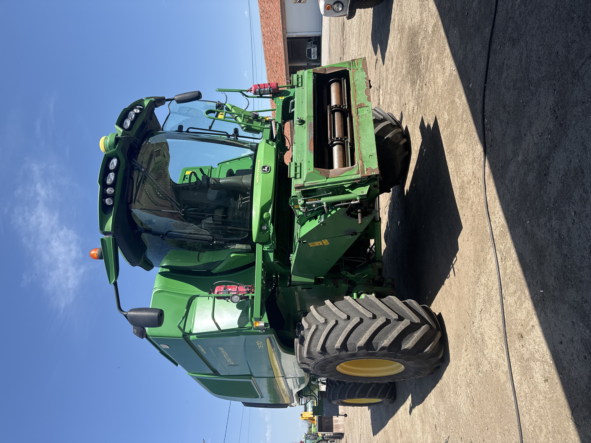 The image shows a green John Deere 7610i self-propelled forage harvester on a concrete surface. The machine is positioned diagonally in the frame. The harvester's front cutting unit is visible. The sky is blue. A building with a red tile roof is partially visible in the background. A power line runs across the concrete surface. Another piece of agricultural equipment is visible in the background. The location is Sarnevo, Bulgaria.