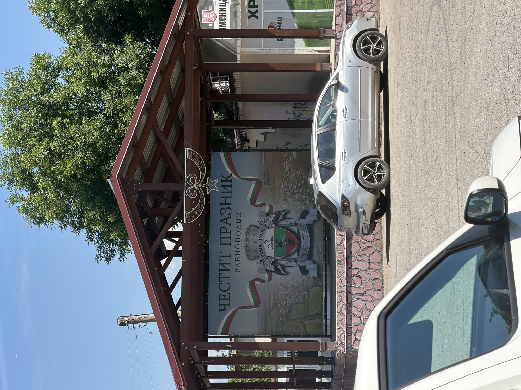 The image captures a street scene in Ravno pole, Bulgaria, on a sunny day. In the foreground, the hood and windshield of a white vehicle are visible, suggesting the photographer is inside or next to another car. A silver hatchback is parked to the right of the white vehicle.
Behind the cars, a large banner is displayed on an open-air structure with a burgundy roof and brown wooden supports. The banner features the text "ЧЕСТИТ ПРАЗНИК" (Happy Holiday) and "РАВНОПОЛЦИ" (Ravno pole residents), along with the coat of arms of Ravno pole, which includes two storks and a shield with a horse.
To the right of the banner structure, a building with windows is visible, with signs that are partially obscured but suggest commercial activity. One sign displays the Bulgarian flag colors and text that appears to be a town's name and dates. Another sign shows the Bulgarian word "ХРАНИЦА" (grocery store). The ground is paved with asphalt, and a low stone wall, decorated with pink and beige painted stones, runs along the base of the banner structure. Green trees are visible in the background, and the sky is clear blue with a faint airplane trail.