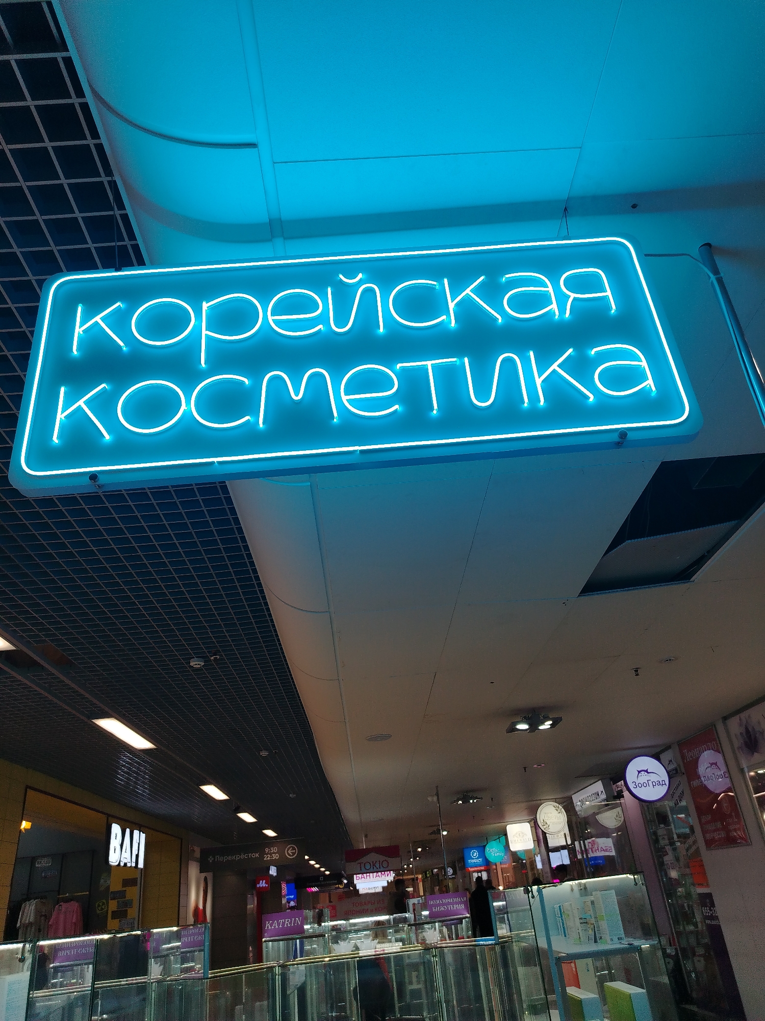 The image shows a view inside a shopping mall in Izhevsk, Russia. The dominant element is a bright blue neon sign that reads "корейская косметика" (Korean cosmetics) in Russian. Below the sign, several shops and boutiques are visible. There is a store named "BAFI" on the left, and signage for "Перекрёсток" (a grocery store chain), "TOKIO," and "KATRIN" can be seen in the background. Also visible is a sign for a shop called "ЗооГрад". A blue ceiling with a grid pattern is visible above the sign. Glass display cases filled with items are in the foreground. People are visible walking through the mall.