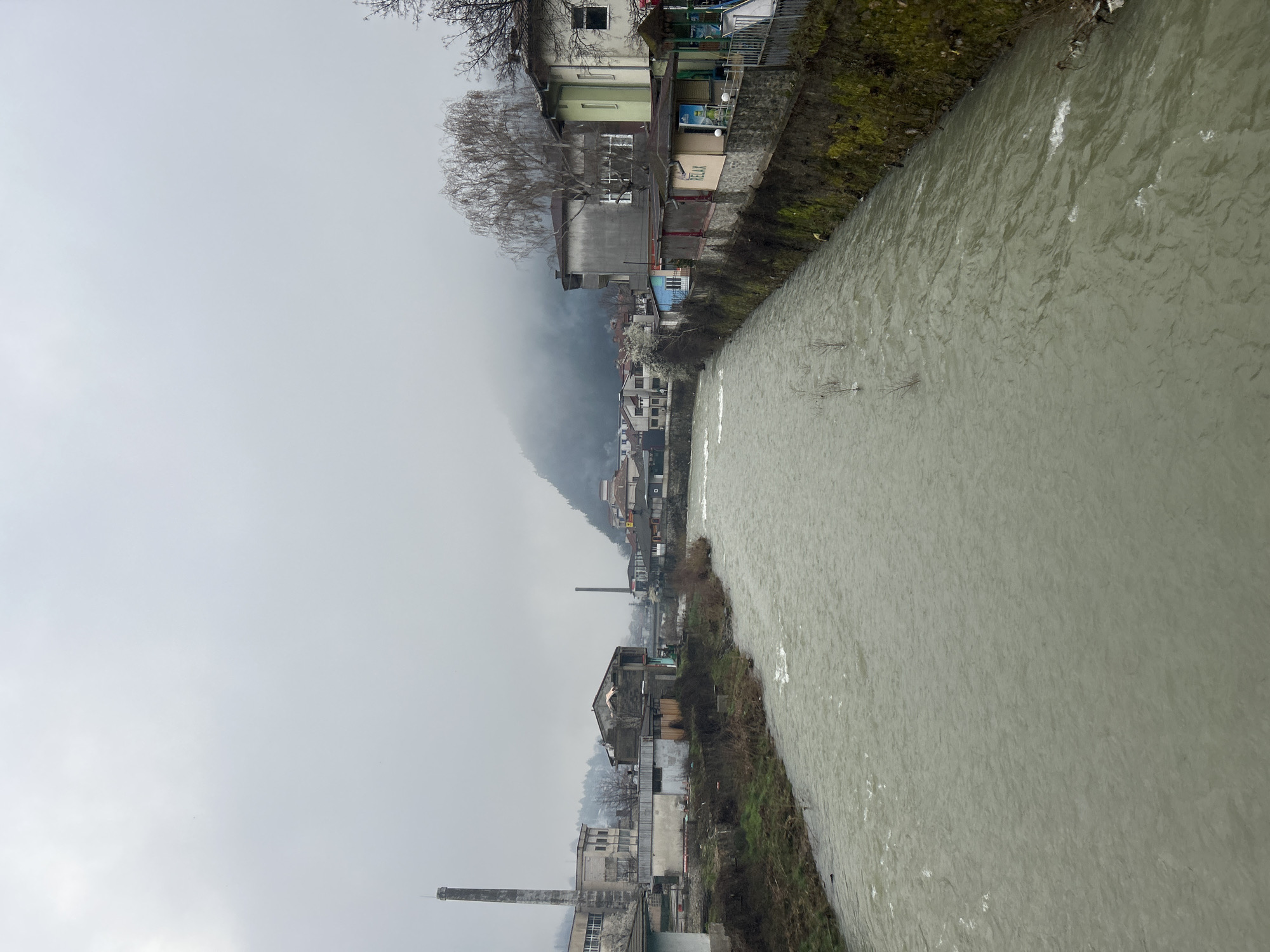 The image shows a river flowing through a city, likely in Zlatograd, Bulgaria. The riverbank is lined with buildings, including residential houses and industrial structures such as a factory with a smokestack. A mountain is visible in the background, partially obscured by fog or mist. The sky is overcast and gray. The river water is murky and moving quickly. The buildings along the riverbank vary in age and condition.