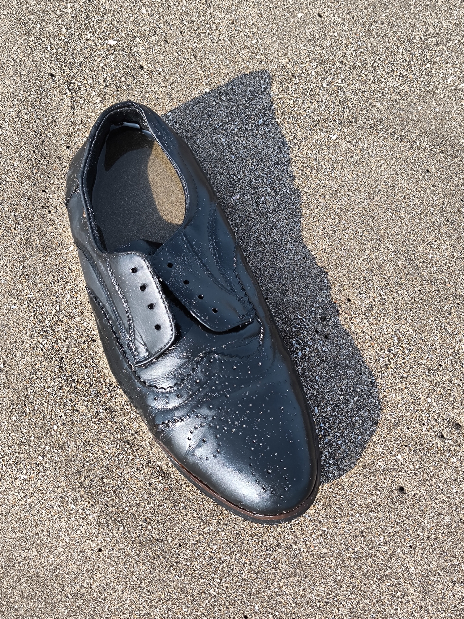 A single, dark-colored dress shoe lies on a sand-covered surface. The shoe is oriented diagonally in the frame, with its toe pointing downwards. It appears to be without laces. The surface beneath the shoe is sand. Based on information available about Bali and the fact the image originated from Canggu, Indonesia, the sand is located in Canggu, Indonesia.