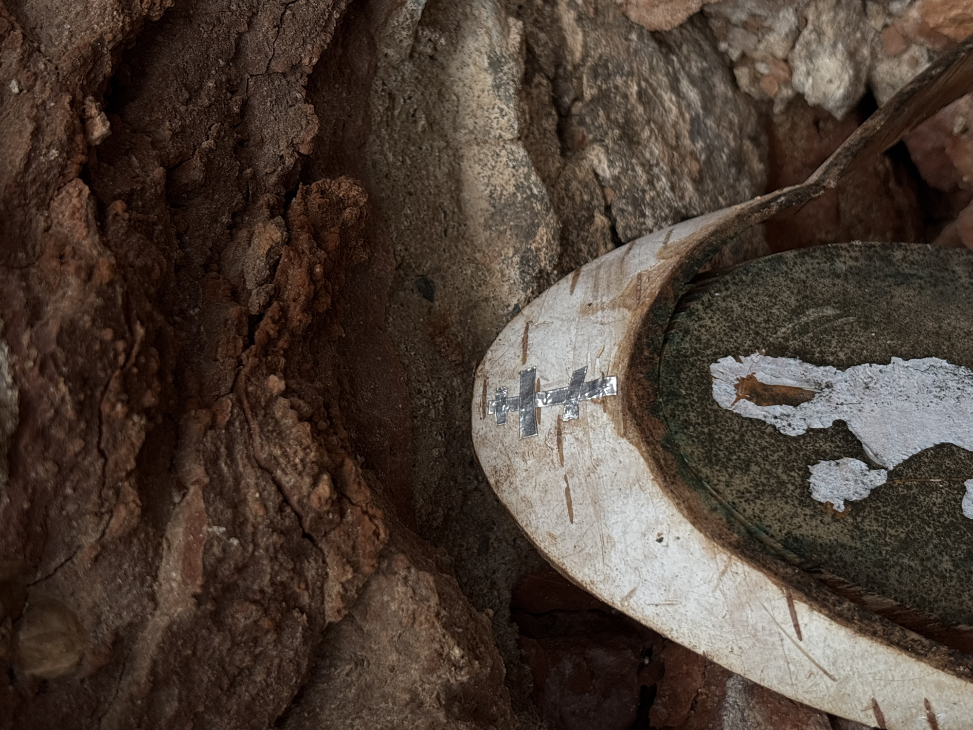 An extreme close-up shot captures a portion of an old, circular object nestled amidst rough, textured rock formations. The object, possibly a lid or part of a vessel, has a worn, light-colored rim, appearing off-white or pale brown, with some vertical scratches or marks. On this rim, a distinct cross-like symbol is visible, formed from several small, reflective, rectangular pieces, possibly metal or tape, suggesting either a repair or a decorative/symbolic addition.
The interior of the circular object is a dark, speckled surface, from which patches of white material are flaking off, revealing the darker layer beneath. Extending from the upper right corner of the frame, a thin, dark, curved piece of material, possibly wood or metal, appears to be part of or associated with the main object, nearly touching its rim.
The background is dominated by highly textured, earthy or rocky surfaces, primarily in shades of dark brown, reddish-brown, and some lighter greyish-brown tones. These surfaces exhibit deep cracks, rough edges, and irregular contours, indicating a natural or rustic setting. No discernible activity or event is taking place, presenting a static, detailed view of the object and its immediate surroundings. The lighting suggests daytime, but without direct sunlight, leaving the time of day and weather indeterminate. No legible text is visible in the image. Given the location context of Staroe Rakomo, Russia, the object could be a historical or archaeological artifact found in a natural or ancient site.