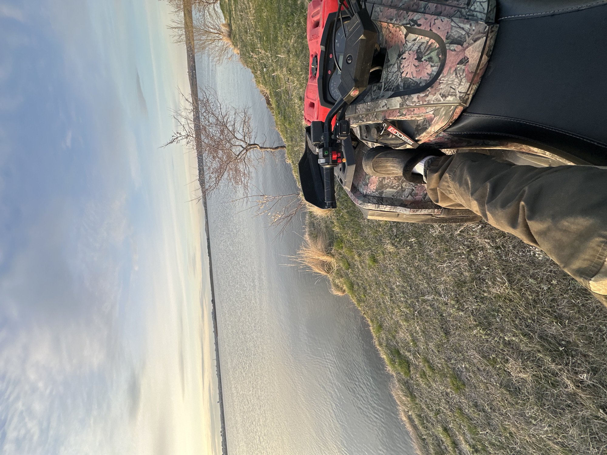 The photograph shows an all-terrain vehicle (ATV) positioned at the edge of a body of water. The ATV is predominantly red and features camouflage detailing on some of its surfaces. A leg clad in a brown garment is visible, suggesting a person is operating or sitting on the ATV. The ATV is parked on grassy terrain. In the background, the body of water extends towards the horizon, reflecting the sky. The sky is a pale blue with light clouds. Trees are sparsely scattered along the shoreline. The photo appears to be taken during the late afternoon or early morning, based on the lighting. The location is an unnamed city.
