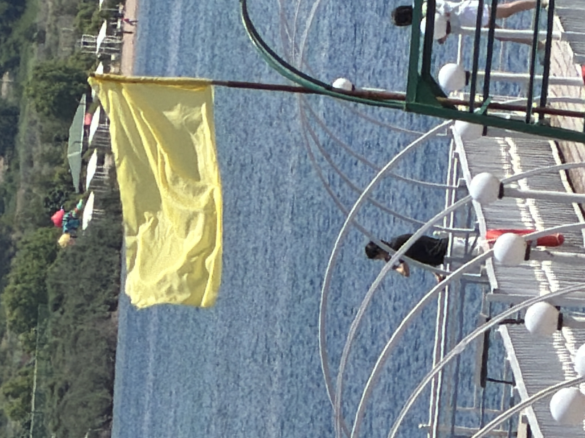 A bright yellow flag billows in the wind on a pole against a backdrop of blue water. In the foreground, a man in a black shirt stands on a white railinged walkway, looking to his left. To the right, a person in a white outfit stands on the same walkway, facing away from the camera. The walkway is adorned with spherical white lights. In the distance, a beach is visible with people and umbrellas, backed by green foliage and buildings.