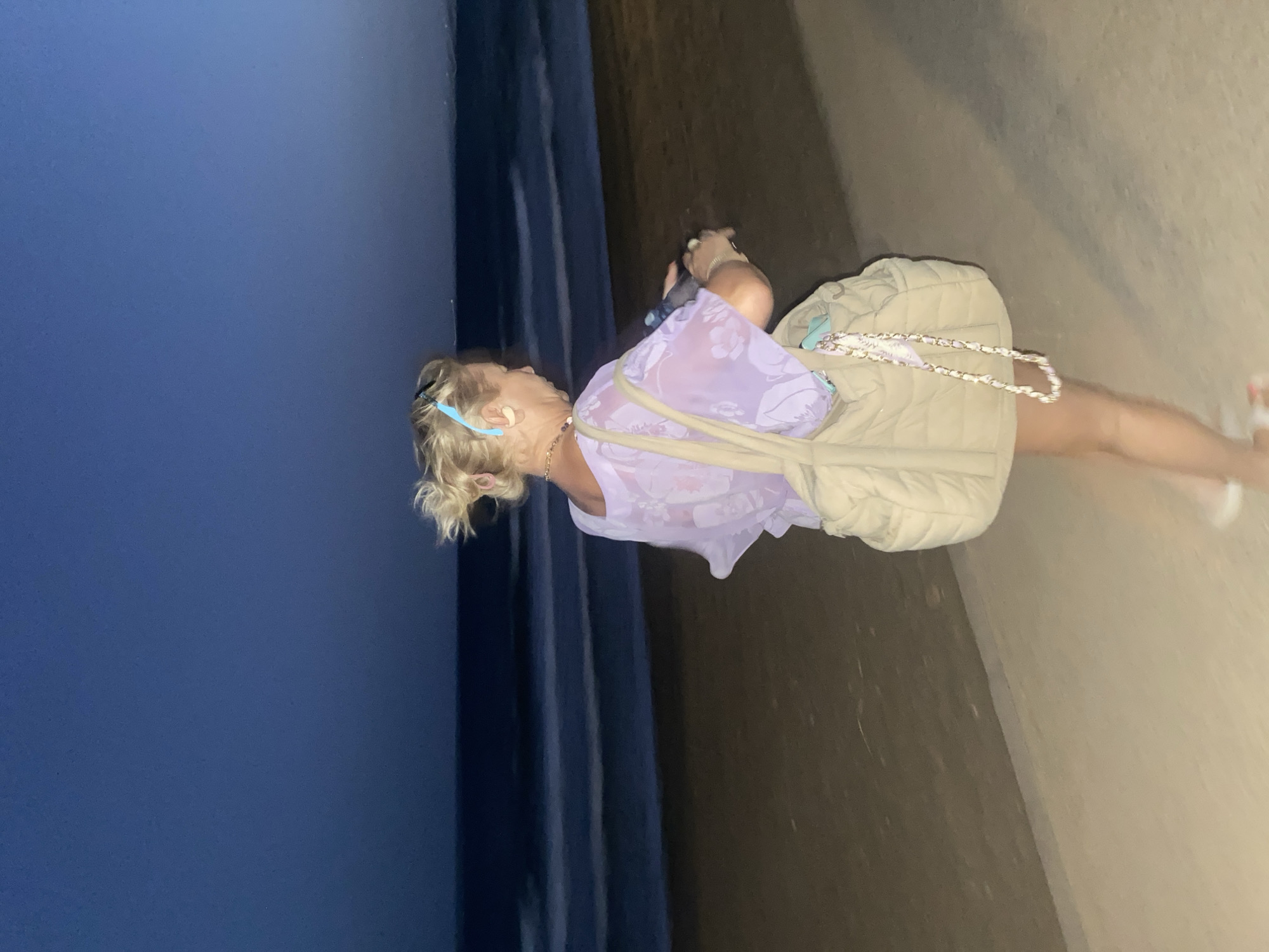 A woman with blonde hair pulled back in a bun and wearing sunglasses is standing on a beach promenade at dusk. She is wearing a light purple, floral print top and carrying a beige quilted bag with a chain strap. The sea is visible in the background with gentle waves. The sky is a deep blue, transitioning from daylight to night. The image is slightly blurred due to motion. It is a scene of a woman enjoying an evening walk along the beach in Estepona, Spain.