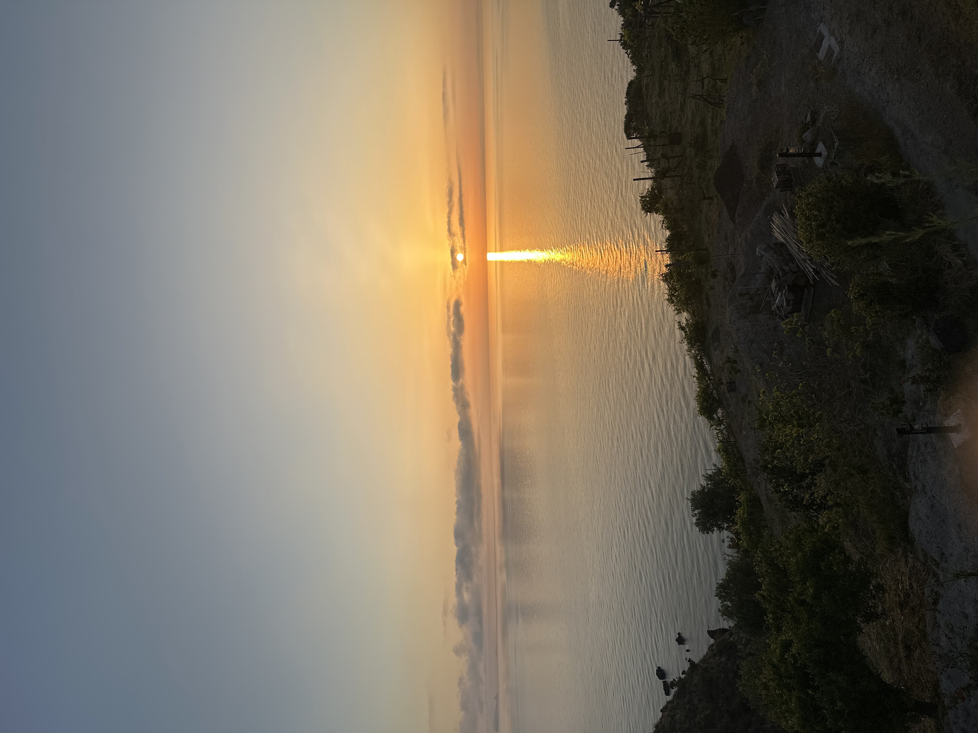 The image captures a serene coastal scene at sunset or sunrise, with the sun low on the horizon over a vast body of water, likely the sea. The sky transitions from a cool blue-grey at the top left to warm orange and yellow hues closer to the sun. The sun itself is a brilliant orange disc, partially obscured by a thin, horizontal cloud formation that stretches across the horizon.
A prominent, bright golden sun pillar reflects intensely on the water's surface, creating a shimmering path directly from the sun towards the viewer. The water is calm with gentle ripples, mirroring the colors of the sky and the sun's reflection.
Along the right side of the image, a dark landmass forms the coastline, appearing to be a rugged hillside or cliff descending to the water. This land is covered with dark green vegetation, including bushes and trees. Hints from cropped views suggest human presence on this land, with visible wooden posts, possibly a fence or part of a structure, and what looks like stacked wood or other materials on disturbed earth. The overall lighting indicates either dawn or dusk, with the warm colors and low sun strongly suggesting sunset. The weather appears clear with only a thin band of clouds near the horizon. No discernible text is visible in the image. The scene conveys a peaceful, natural coastal environment.