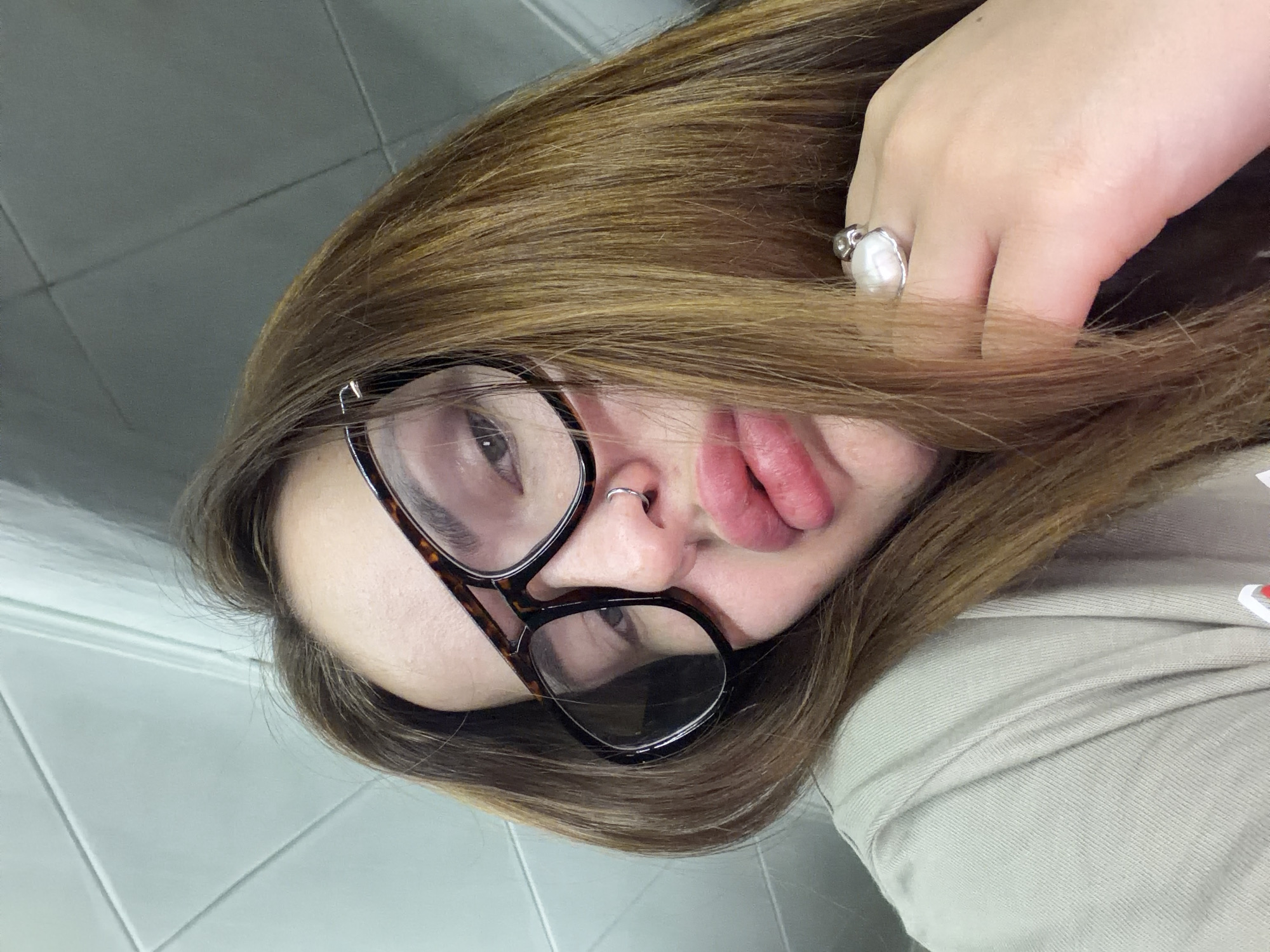 A selfie of a female wearing glasses and a nose ring in Unknown, Unknown. The subject has brown hair and is wearing a ring on her left hand. The background appears to be a tiled wall.