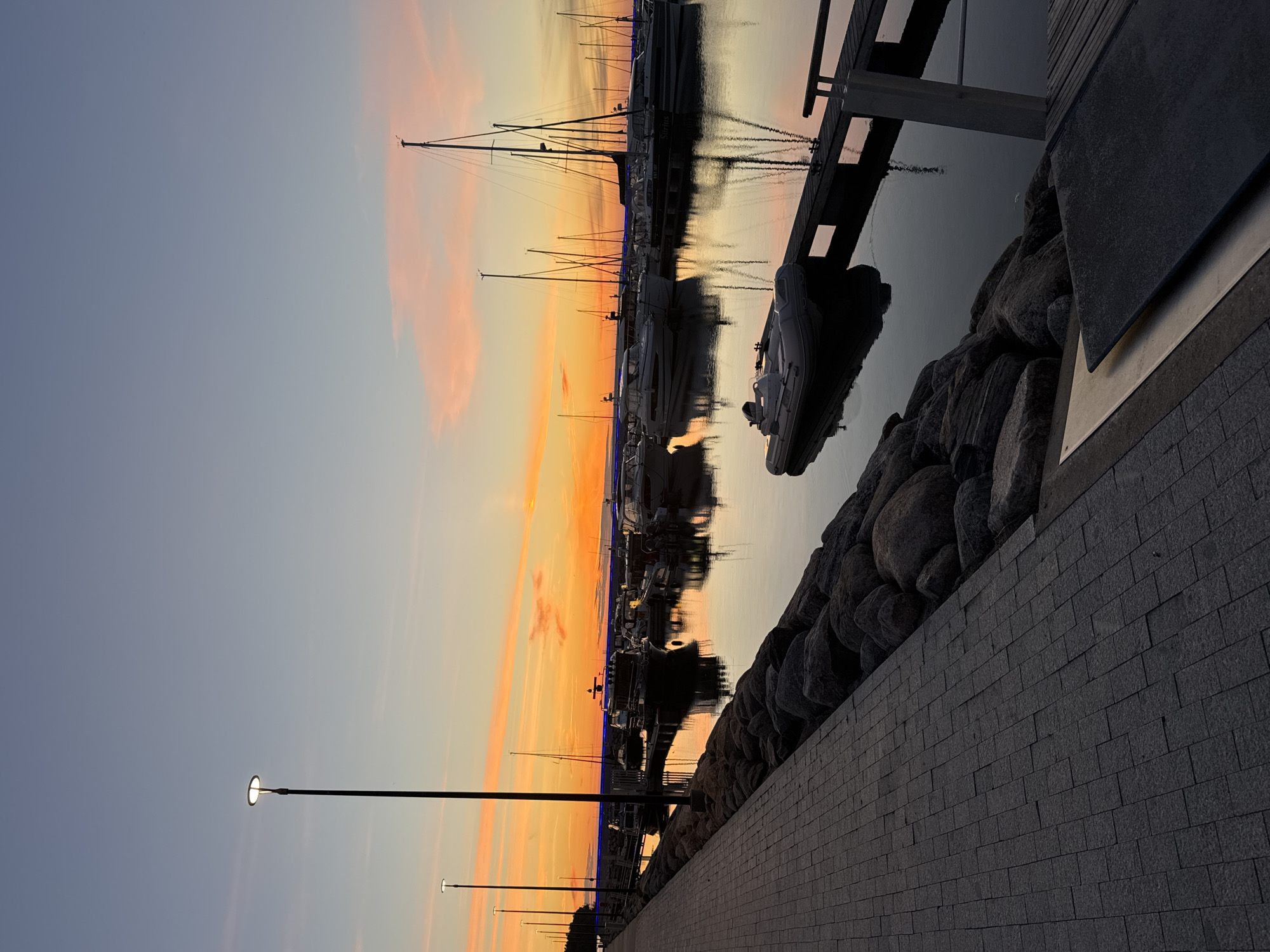 This is a vertical, eye-level shot of a harbor at sunset in Tallinn, Estonia. The sky is a gradient of pale blue at the top, transitioning to orange and pink hues near the horizon, with wispy clouds catching the last light of the sun. A paved walkway runs along the left side of the image, bordered by large, dark rocks. Several lampposts with lit bulbs line the walkway. In the harbor, numerous boats of various sizes, including sailboats and motorboats, are moored. Their reflections are visible on the still water. A small inflatable boat is tied to a wooden pier in the foreground. The overall atmosphere is serene and peaceful, capturing the tranquil beauty of the harbor at dusk.