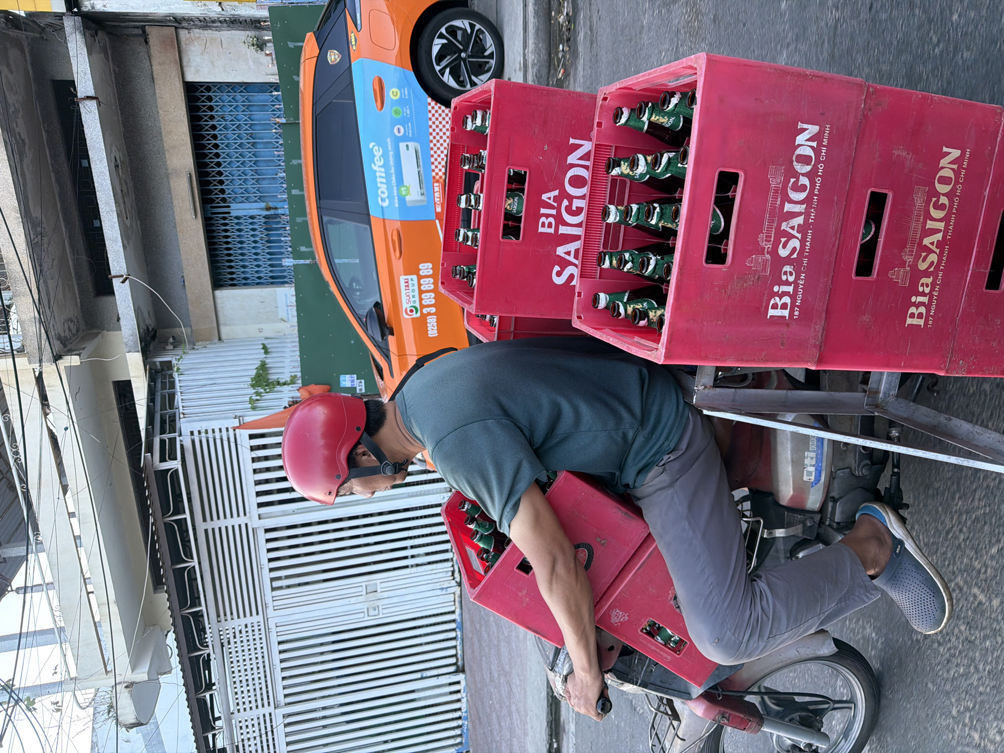 A man wearing a red helmet and a green t-shirt rides a red motorcycle carrying two stacked red plastic crates full of "Bia Saigon" beer bottles. The man is on a paved road in front of a building with a white slatted gate and a parked orange car with "Comfee" and "Suntaxi Group" branding. The scene appears to be in an urban or suburban area in Vietnam, possibly in or near Bac Nha Trang Ward. The lighting suggests it is daytime, likely late afternoon or early morning due to the angle of the shadows.