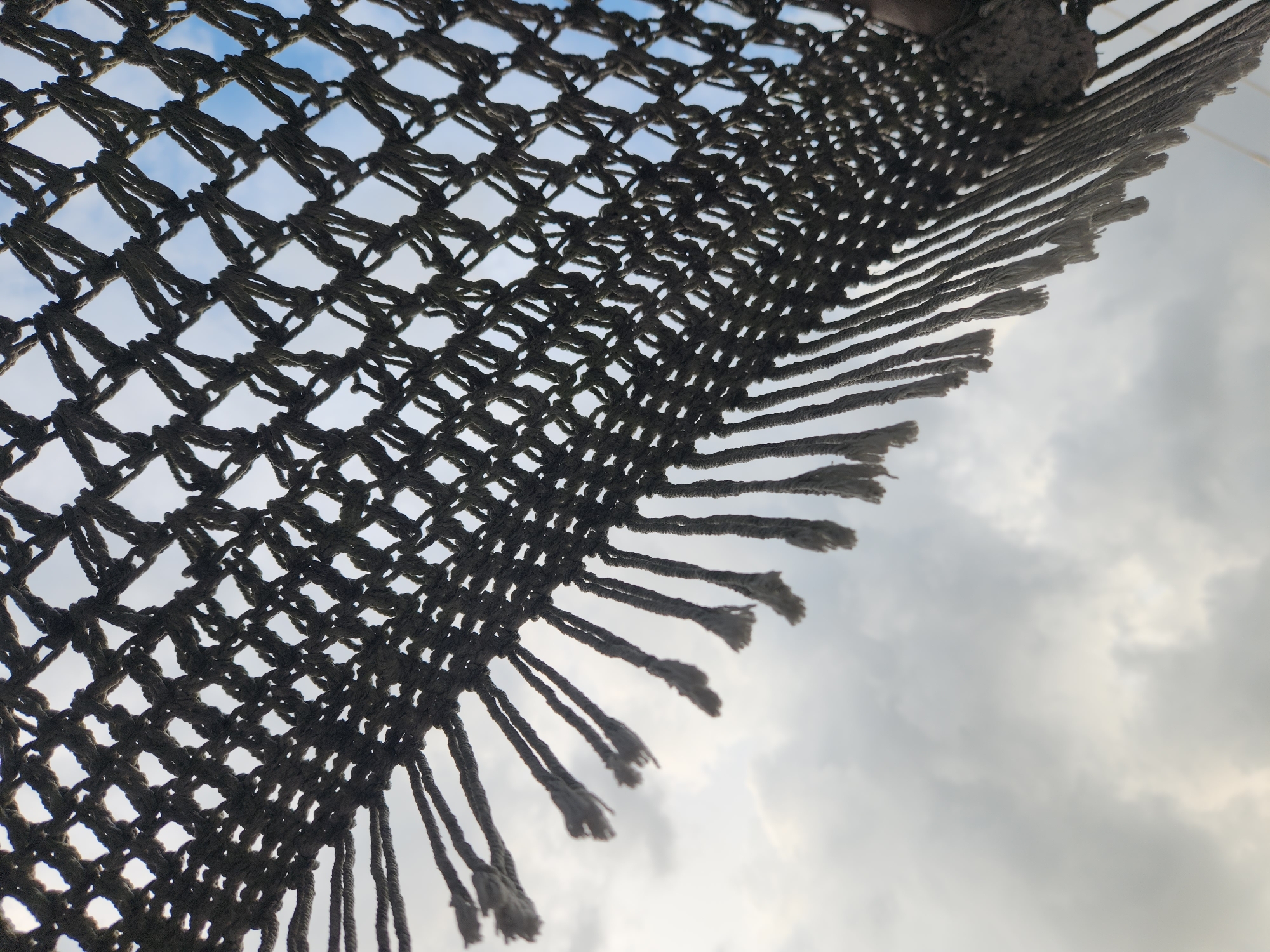 The image shows a close-up view of a thick, dark-colored woven net or fabric, likely part of a hammock or shade structure, with frayed tassels hanging down. The net is viewed from below, with a cloudy sky as the backdrop. The sky is a mix of white and gray clouds, with some hints of blue peeking through. The texture of the woven material is clearly visible, showing individual strands of rope twisted together to form a diamond pattern. The lighting suggests it is daytime, possibly late afternoon or early morning due to the softness of the light. There are no people or animals visible in the image, and no other objects are clearly identifiable. The image is taken outdoors, suggesting a recreational or outdoor setting. There is no information in the image that places it specifically in Maitland, United States of America; this is a detail provided by the prompt that cannot be deduced from the visual information alone.