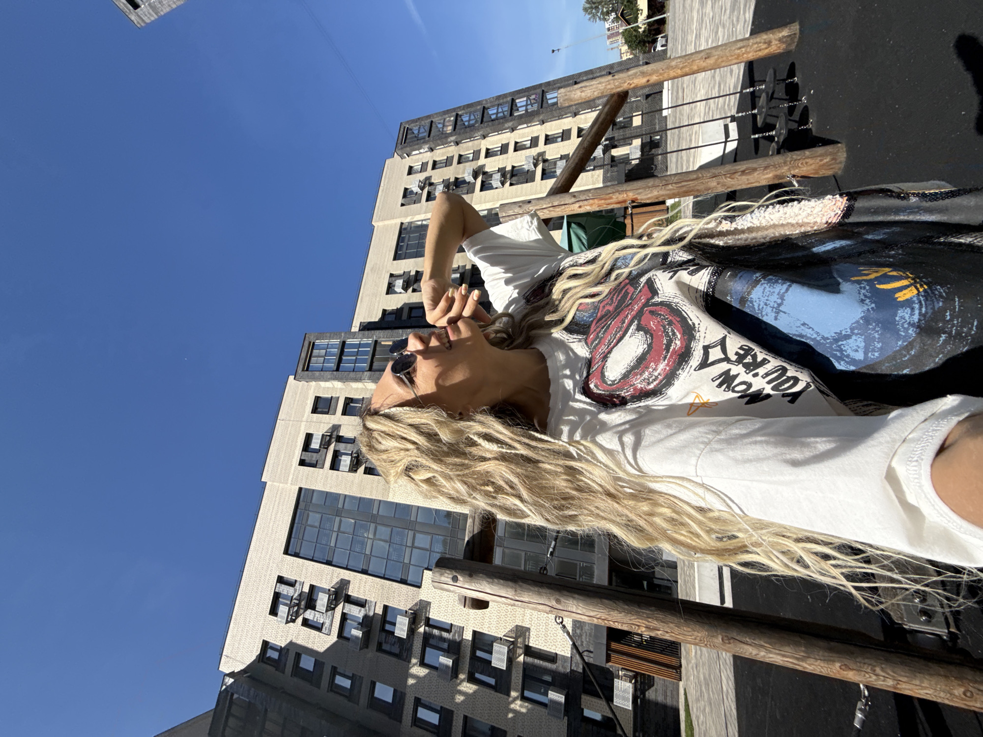 A low-angle, full-shot image captures a blonde woman with long, curly hair in a white t-shirt with a graphic print standing in an outdoor playground. She is wearing sunglasses and has her right arm raised, with her hand near her mouth as she looks upwards. Behind her, a modern, multi-story building with a dark brick facade and lighter colored brick accents dominates the background under a clear blue sky. The playground features wooden support beams and swings. The location is the city of Rusanovo, Russia.